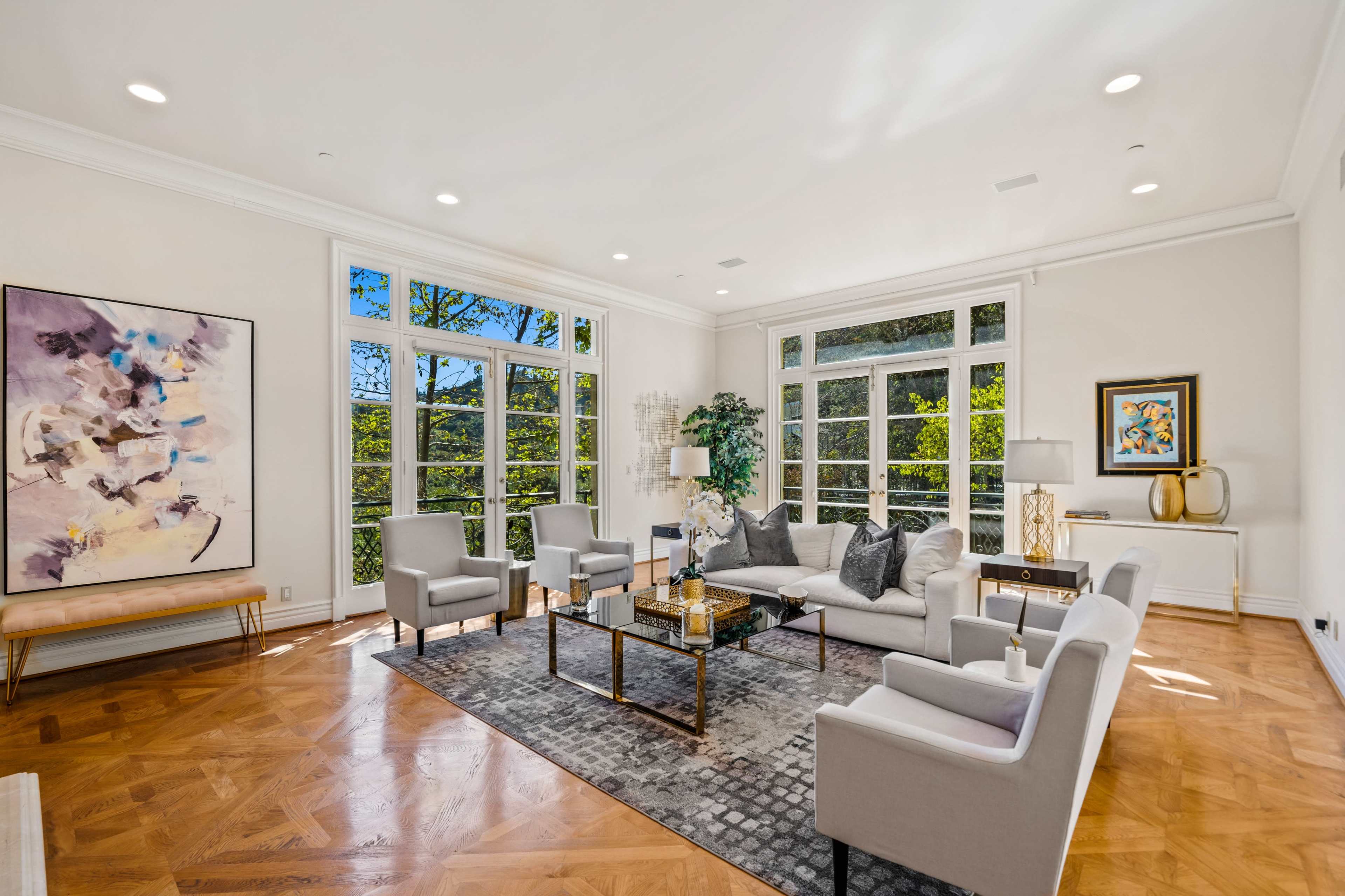 A bright living room features large windows with views of greenery, stylish furniture, and decorative artwork.