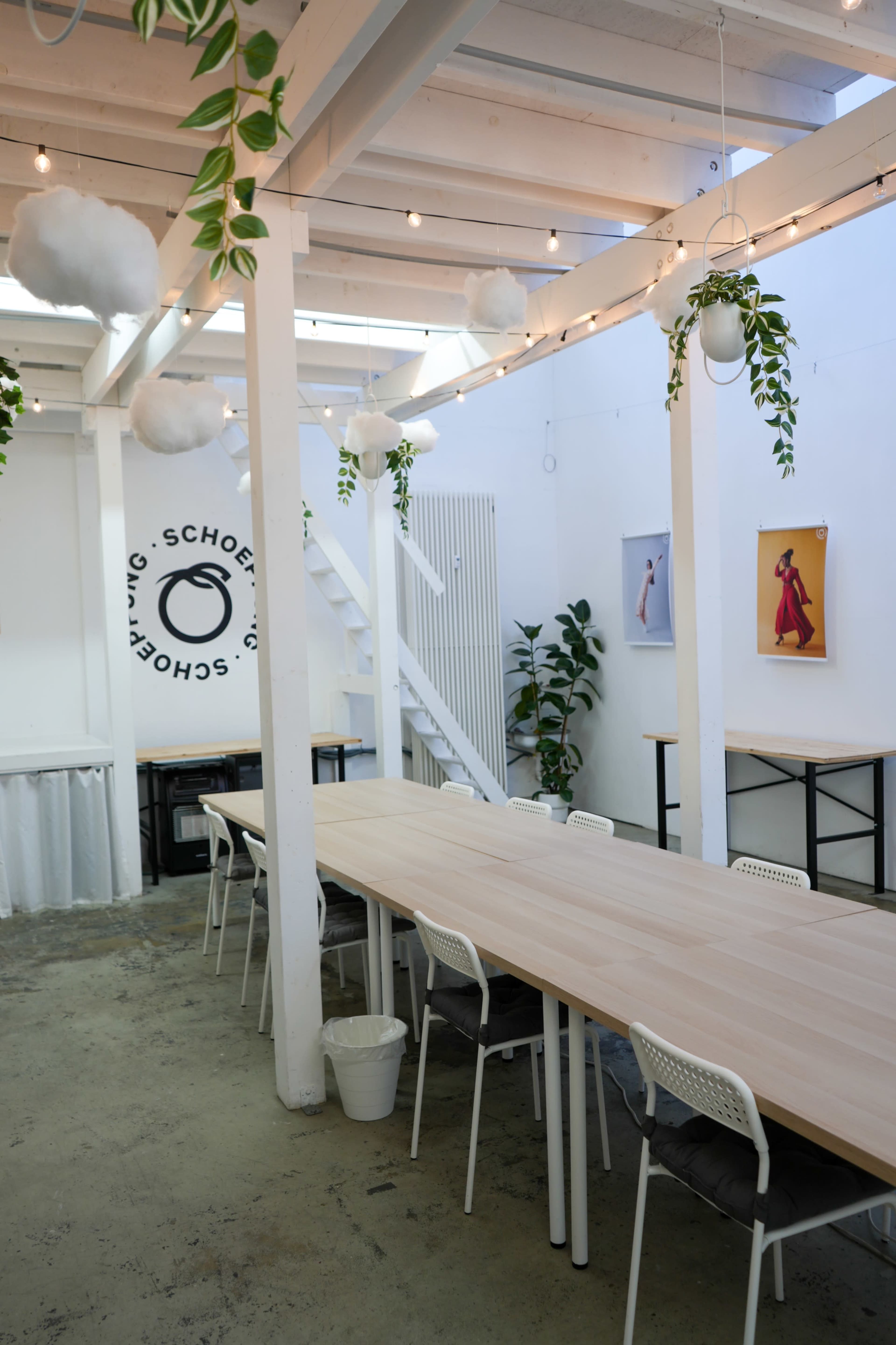 A modern, well-lit interior space featuring a long wooden table, white chairs, potted plants, and decorative hanging clouds.