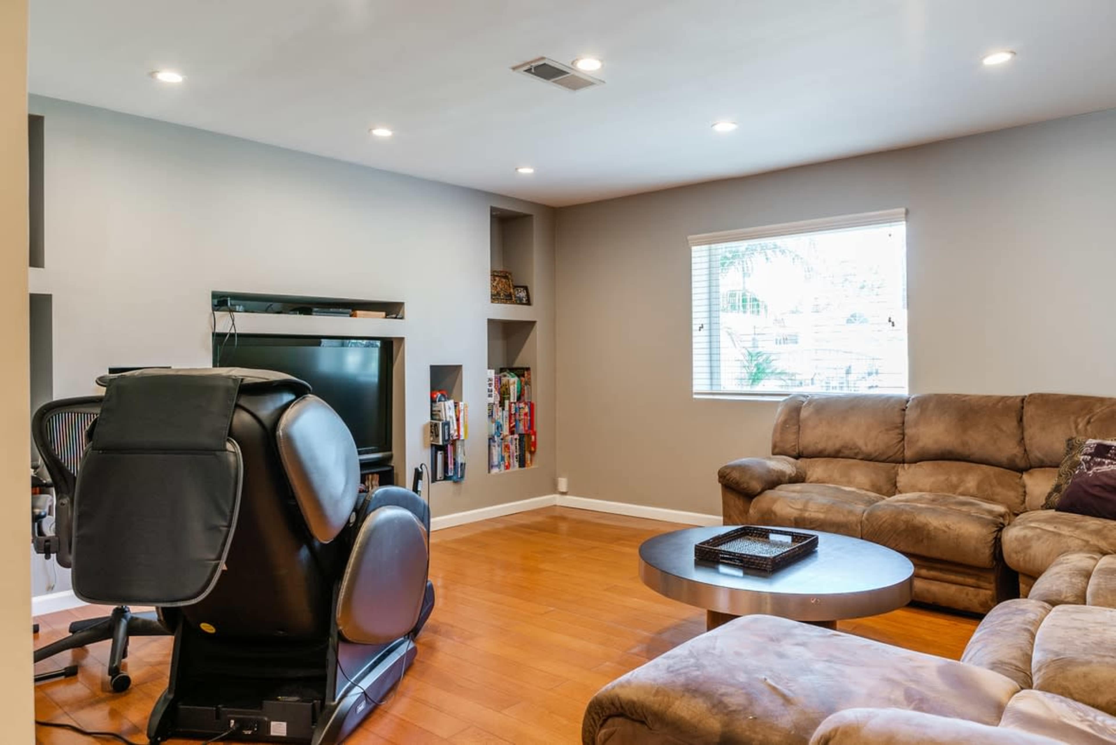The room features a brown sectional sofa, a round coffee table, a large television, and a recliner, with shelves displaying various items.