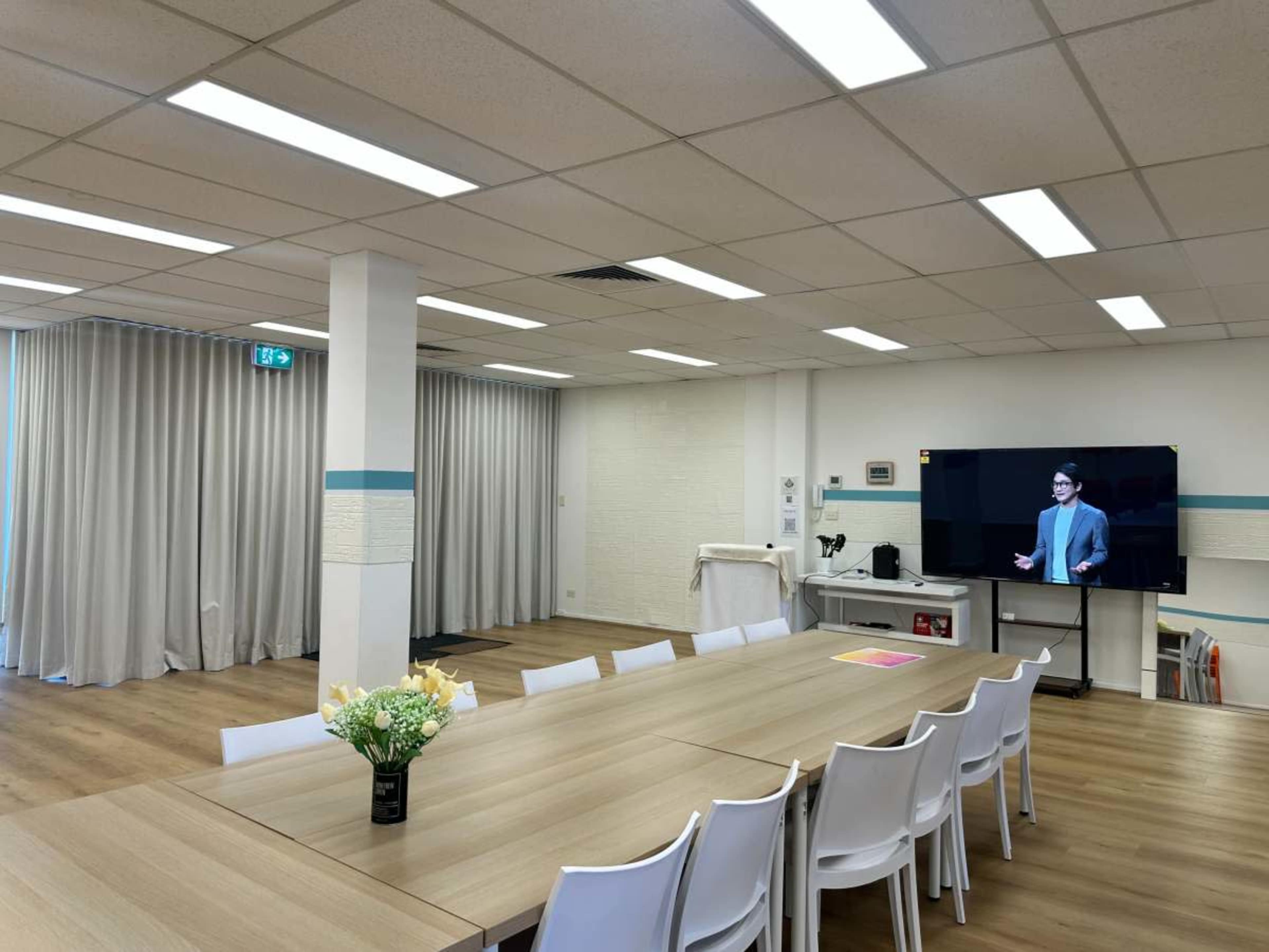 The image shows a modern conference room with a long wooden table, white chairs, a large screen displaying a speaker, and light-colored curtains partially closed.