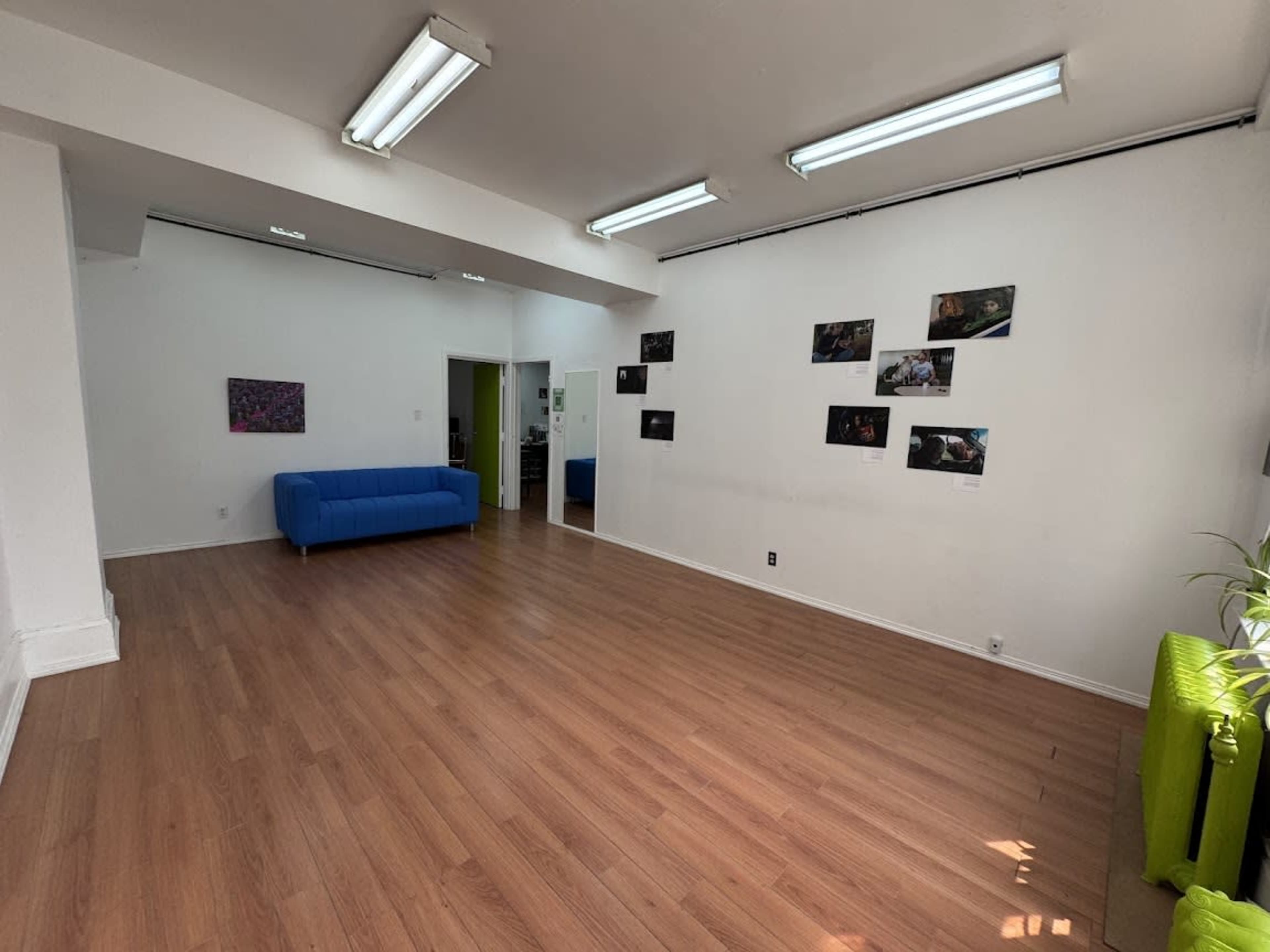 The image shows a spacious, well-lit room with wooden flooring, a blue couch, and several framed photographs hanging on the white walls.