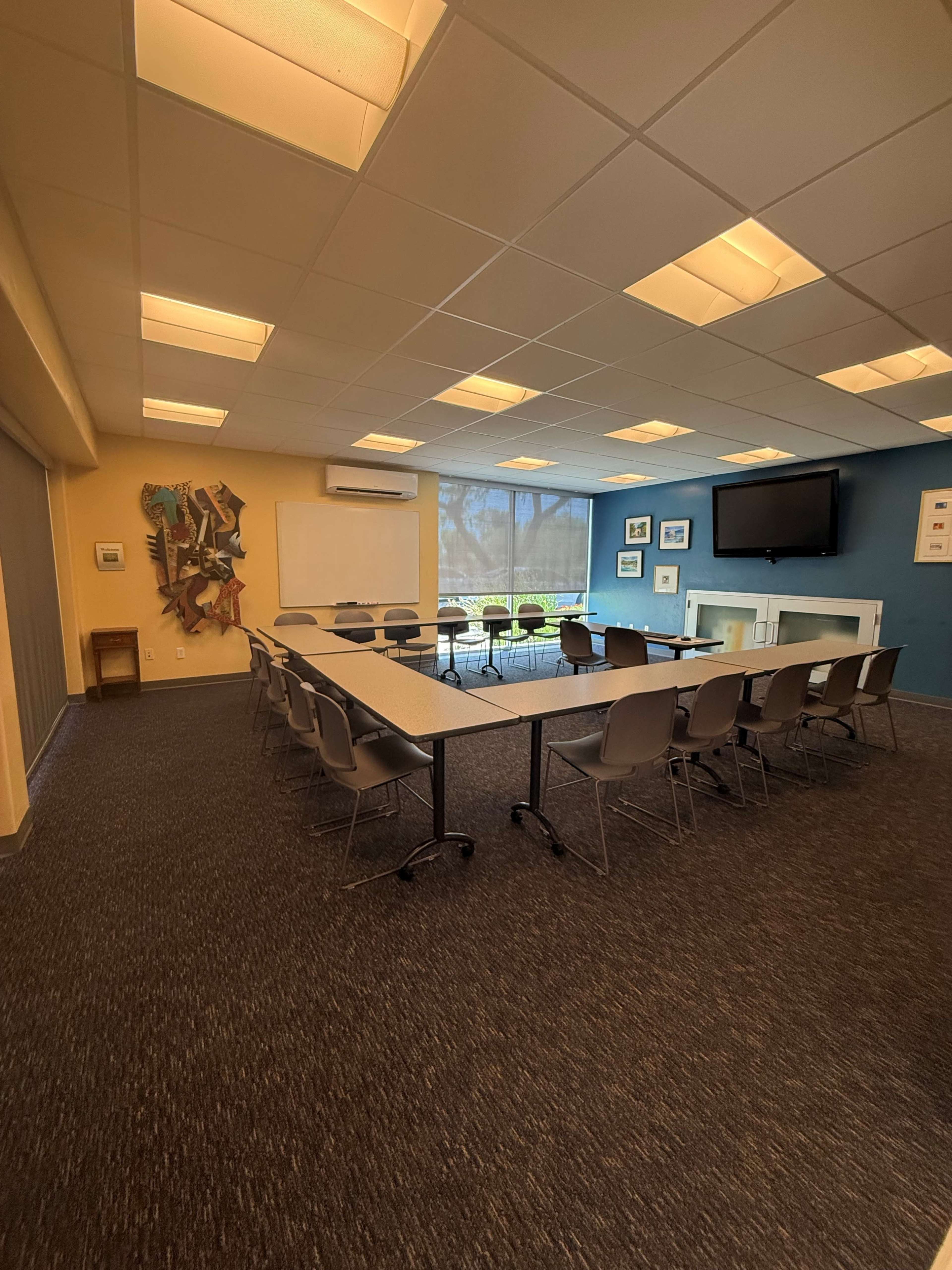The image shows a rectangular meeting room with a long table and chairs arranged for a discussion, featuring a blue and yellow wall, a television, and large windows letting in natural light.