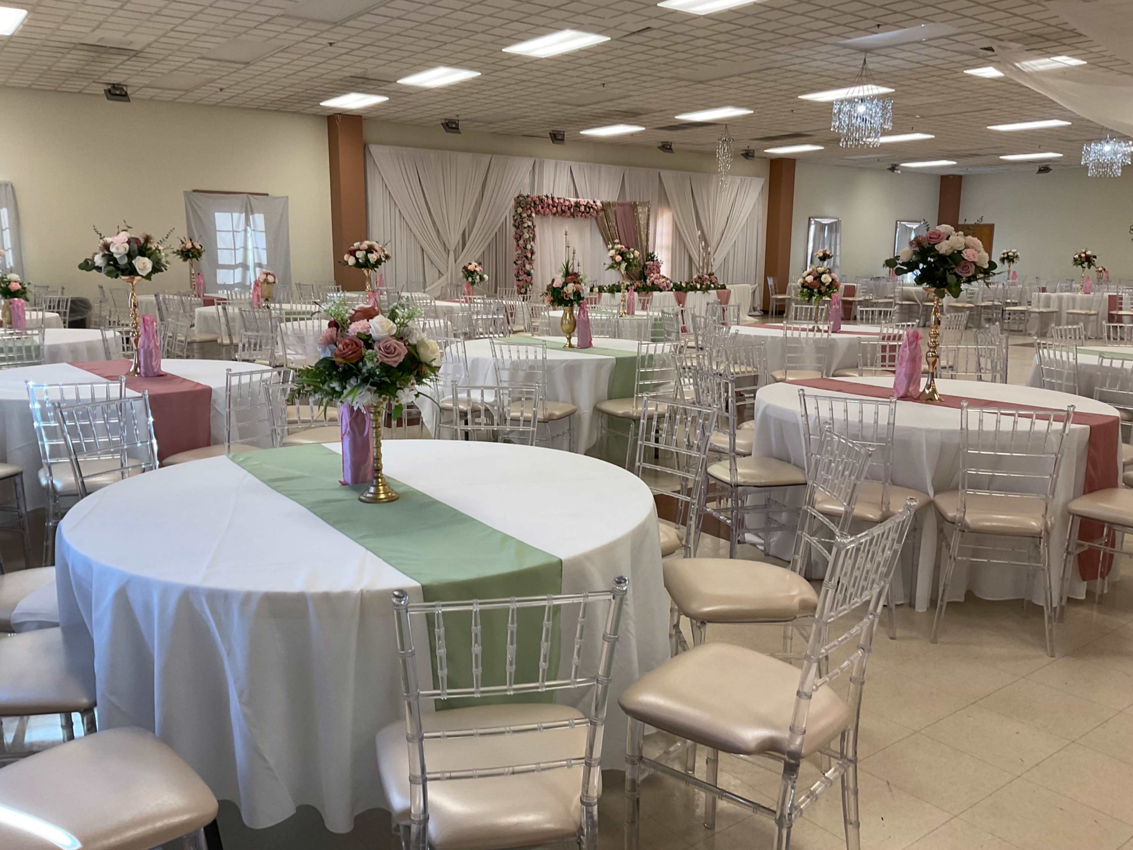 The image shows a decorated banquet hall with circular tables set for an event, featuring floral centerpieces and draped fabric in the background.