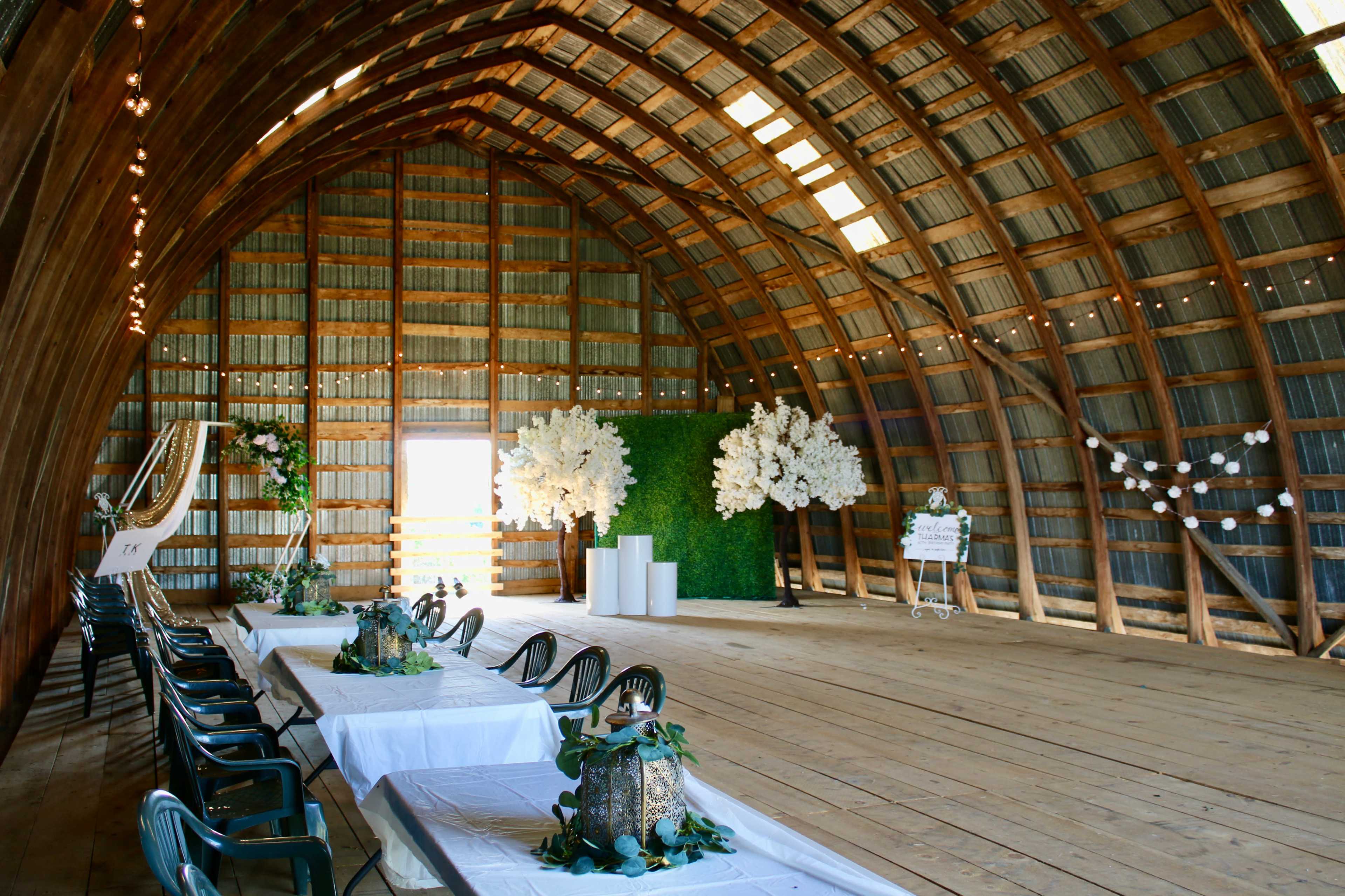 The interior of a rustic barn features a spacious wooden structure with tables arranged along the floor, decorated with greenery and white floral arrangements.