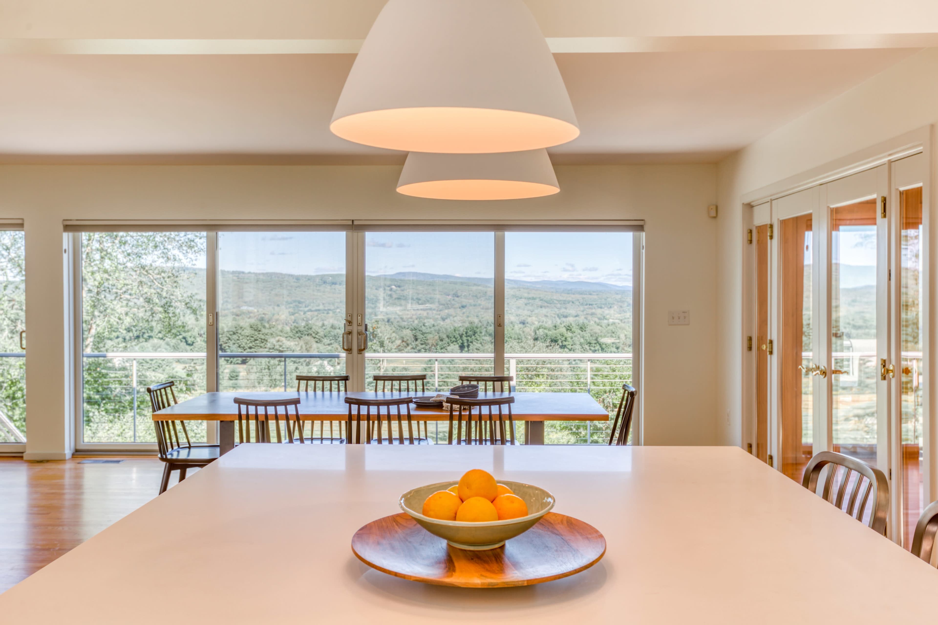 A modern dining area features a white table with a bowl of oranges, large windows offering a view of rolling hills, and a set of wooden chairs arranged around a dining table.