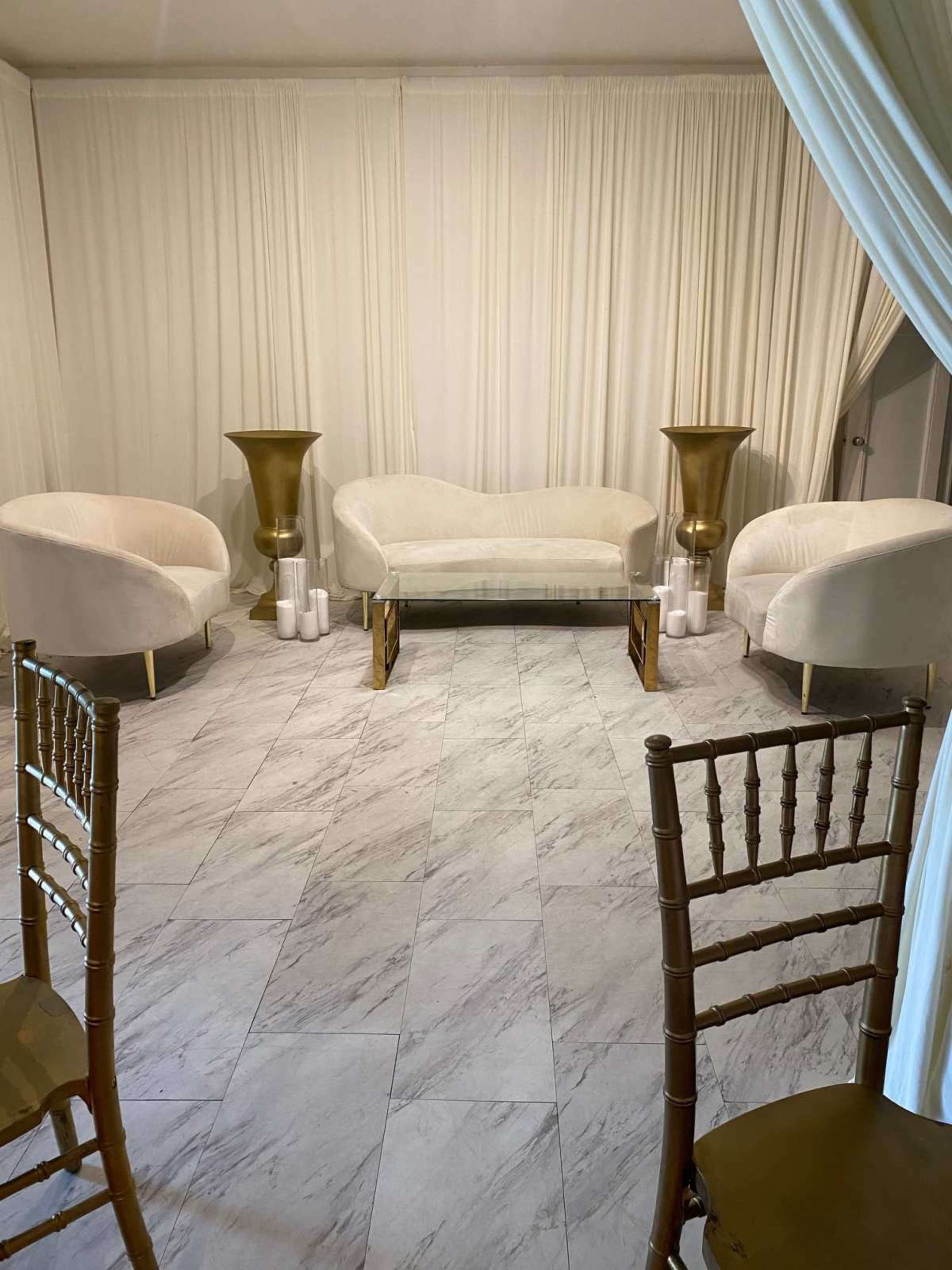 A room features a glass coffee table flanked by two plush white sofas and two gold-accented tall vases, with marble flooring and white drapery.