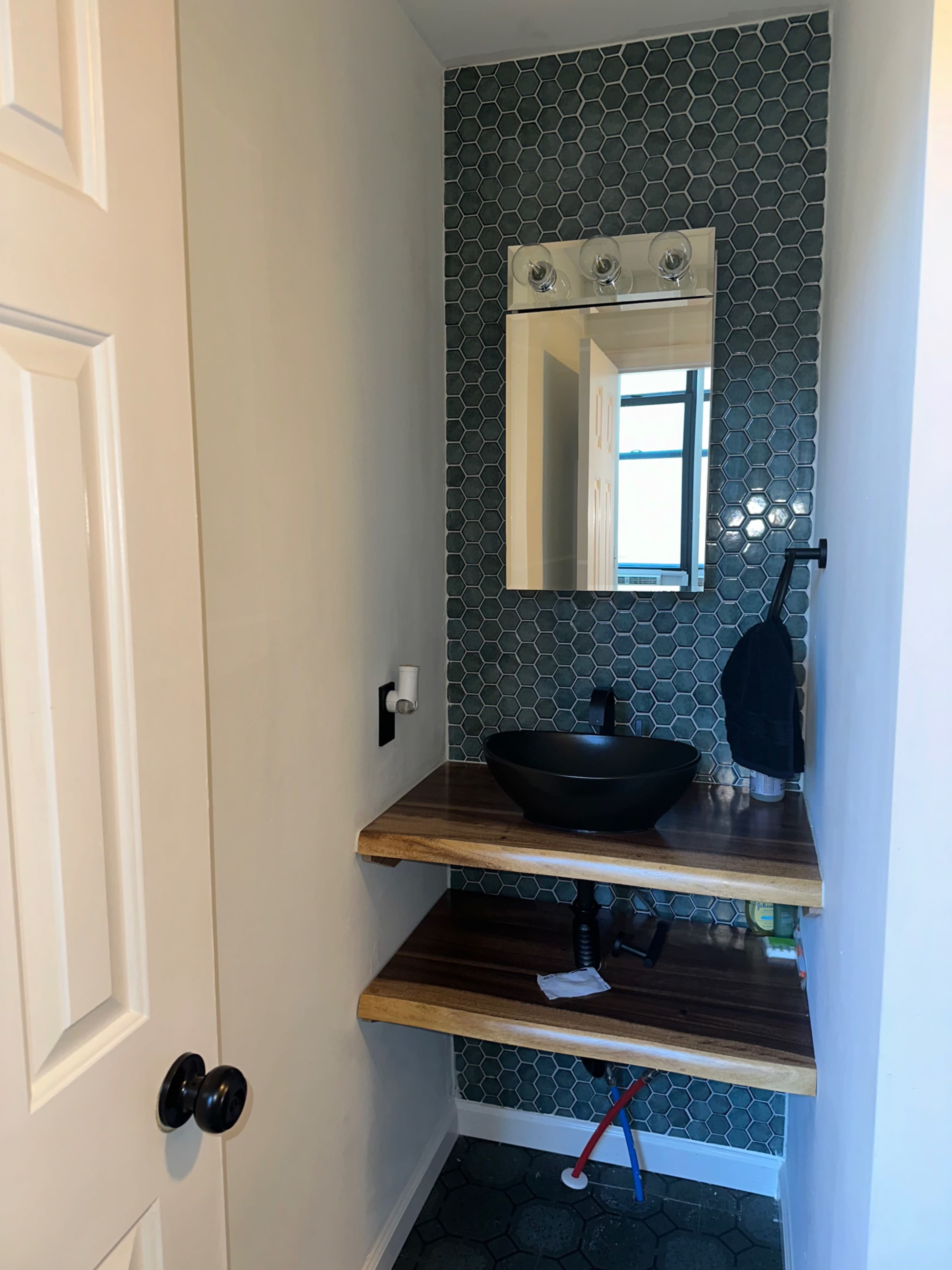 The image shows a small bathroom with a hexagonal tiled wall, a floating wooden shelf, a circular black sink, a mirror with three light fixtures, and a white door.