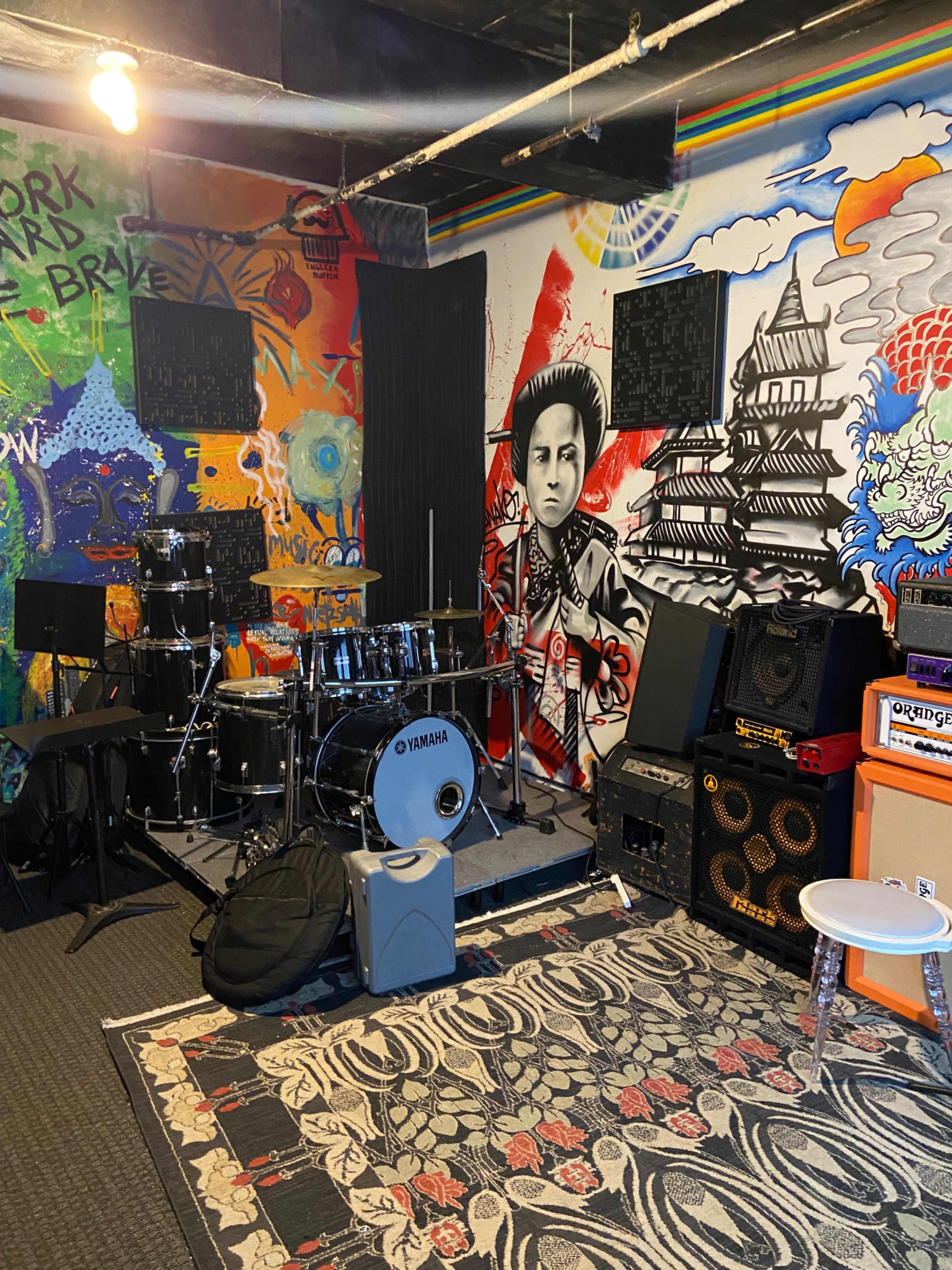 The image shows a music rehearsal space with a drum kit, amplifiers, and a colorful mural featuring a prominent figure and intricate designs on the walls.