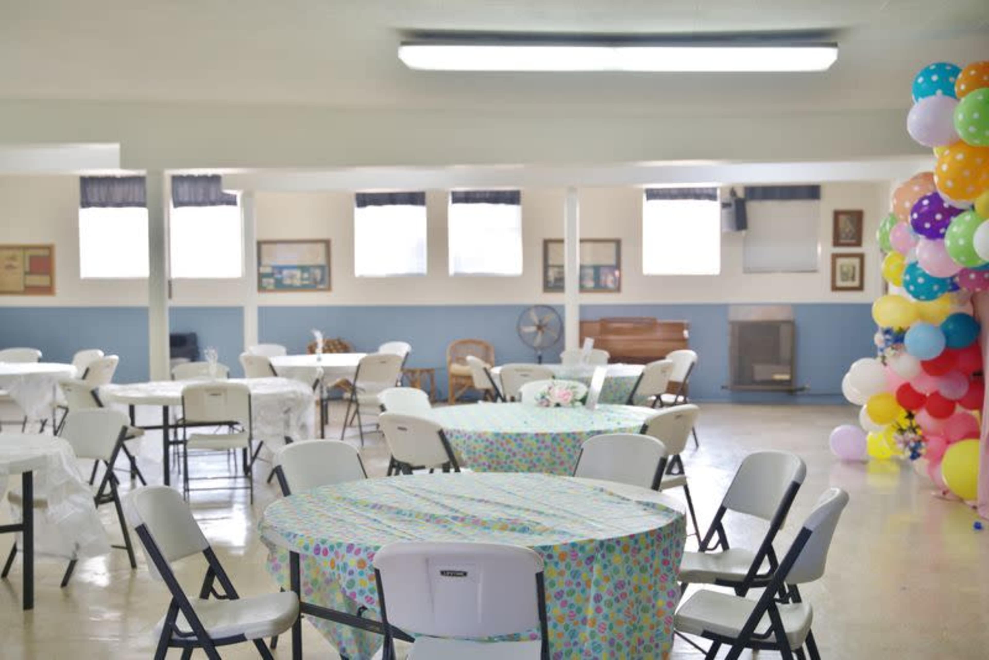 The image shows a spacious community hall with several round tables covered in colorful tablecloths and a large decorative balloon arch in one corner.