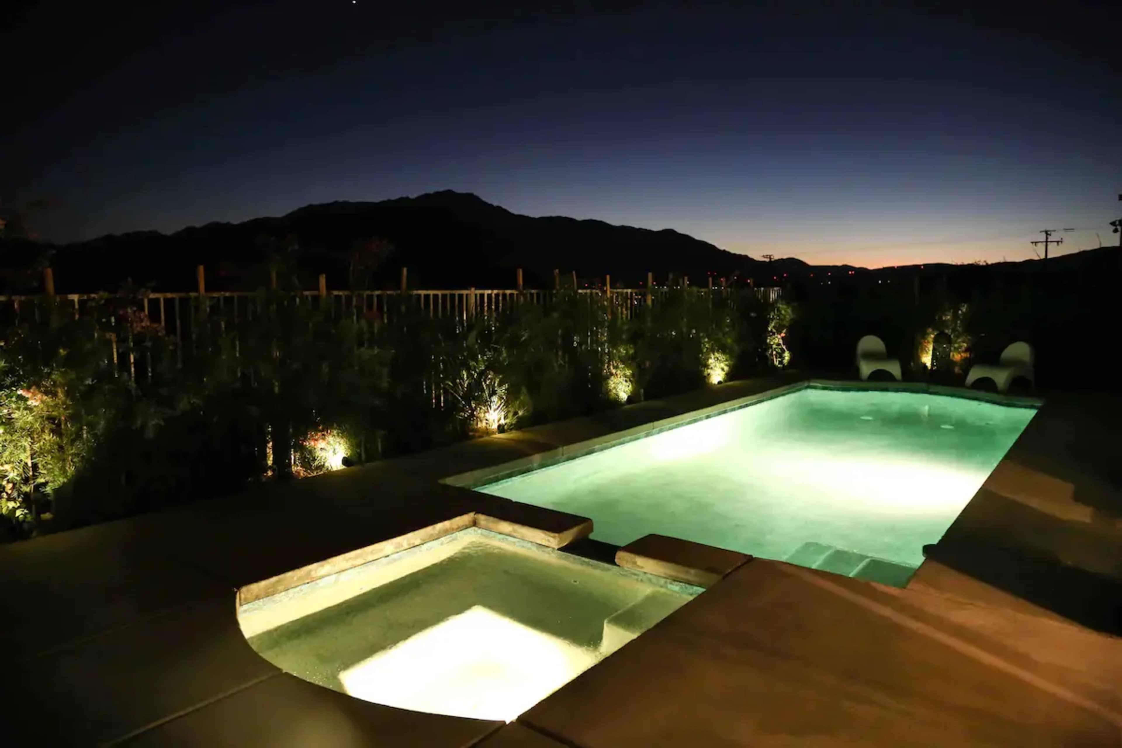 A well-lit pool area is visible at night, featuring a swimming pool and a spa, with mountains in the background and landscaped greenery surrounding the space.