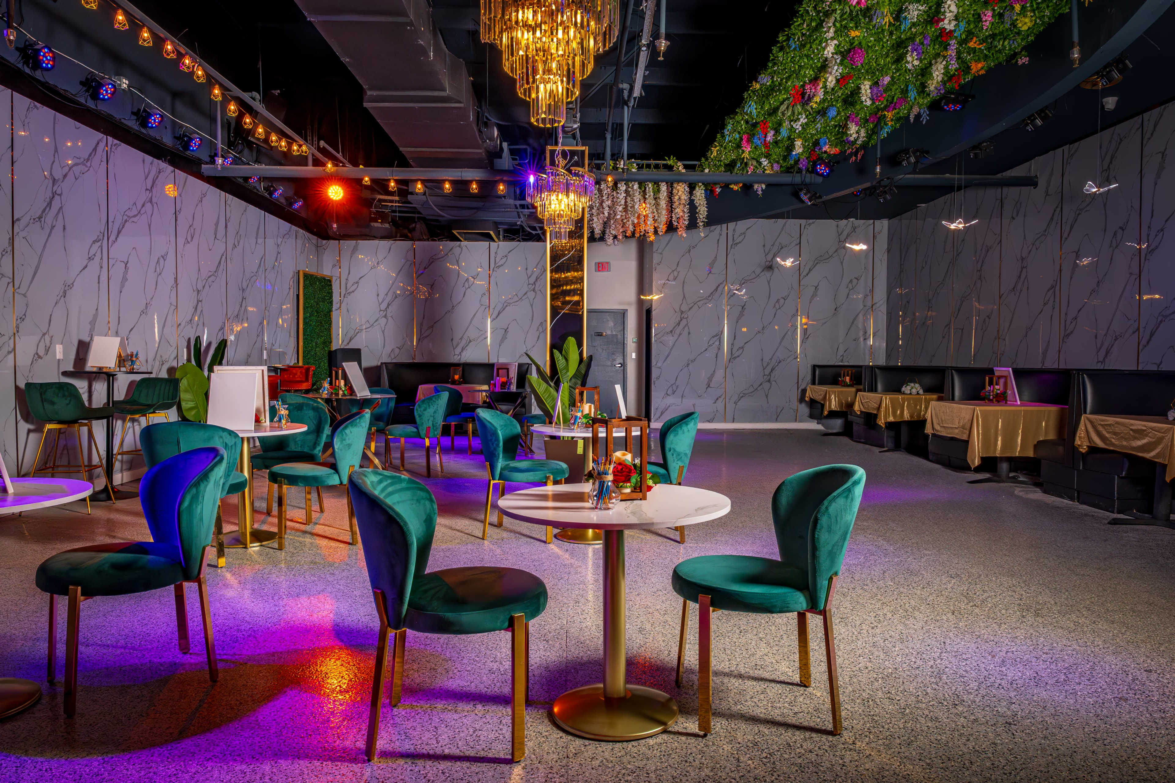 The image displays a modern event space featuring green upholstered chairs, round tables, and decorative lighting under a floral ceiling arrangement.