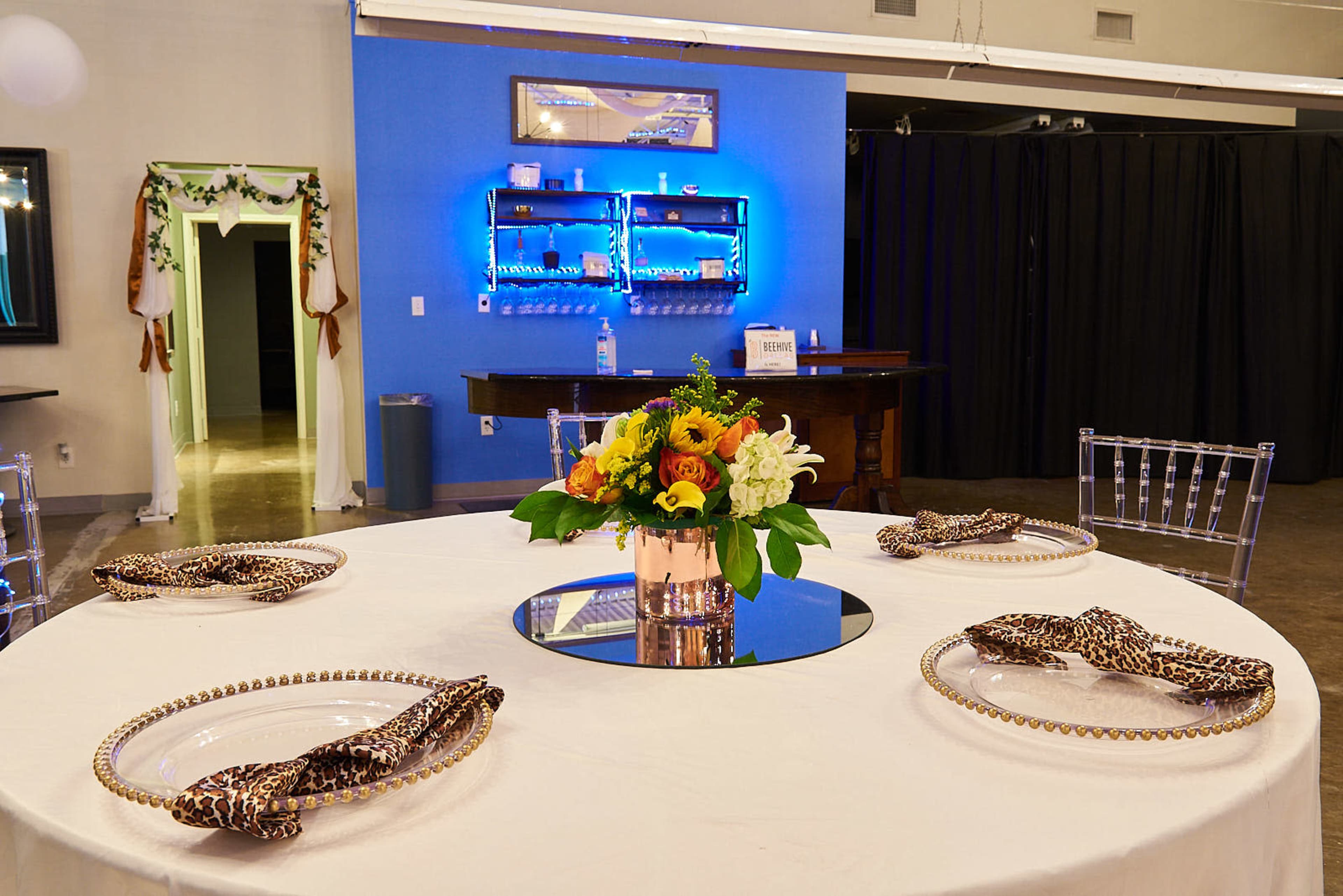 A round dining table is set with elegant plates and a floral centerpiece in a well-lit room featuring blue and dark curtains in the background.