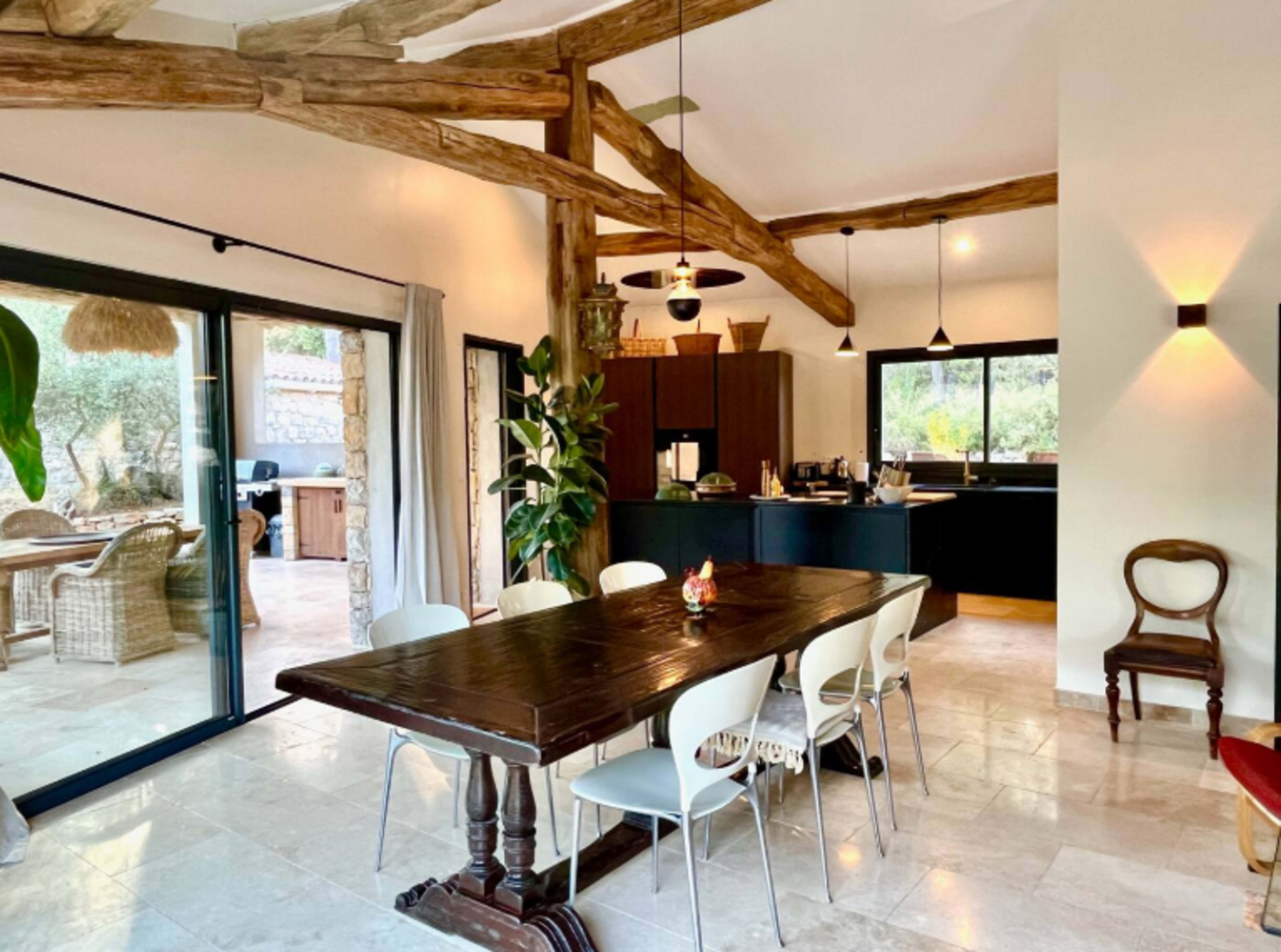 The image shows a spacious dining area with a large wooden table surrounded by chairs, featuring exposed wooden beams and a view into a well-lit kitchen.