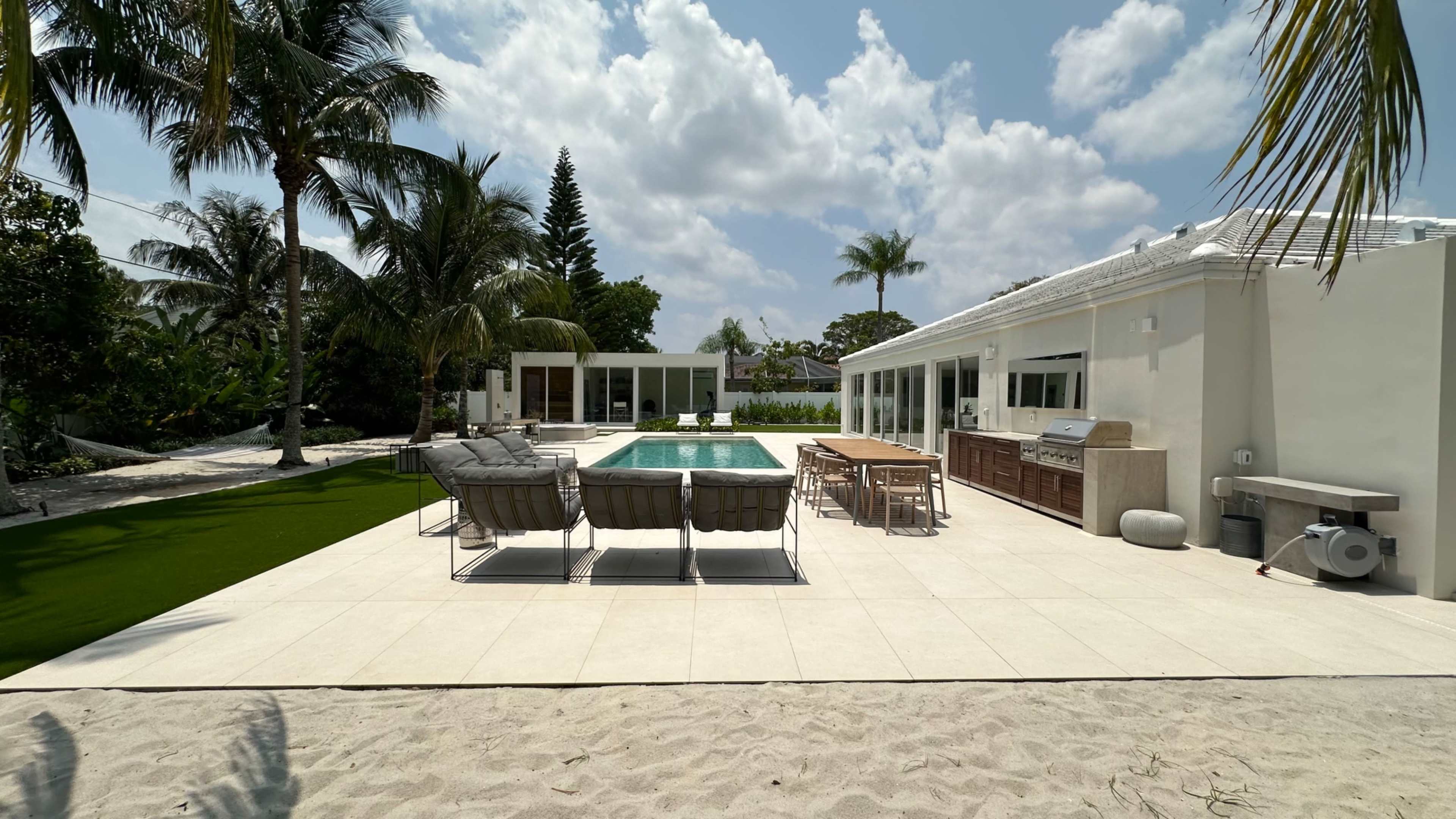 The image shows a modern outdoor space with a swimming pool, lounge chairs, a dining area, and tropical landscaping.