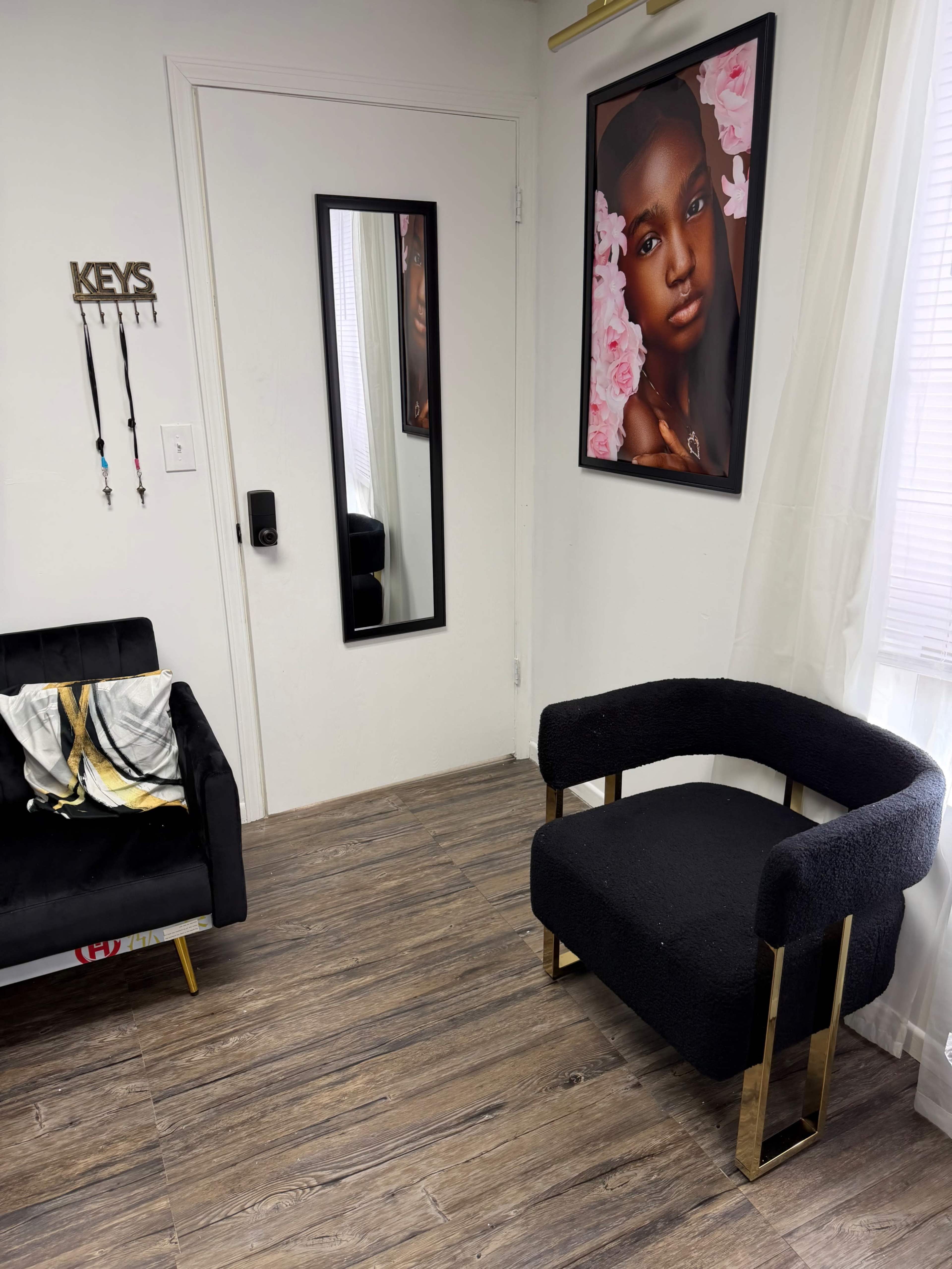 The image features a room with a black armchair and a black sofa, a framed portrait of a woman on the wall, a mirror door, and a key holder mounted beside the door.