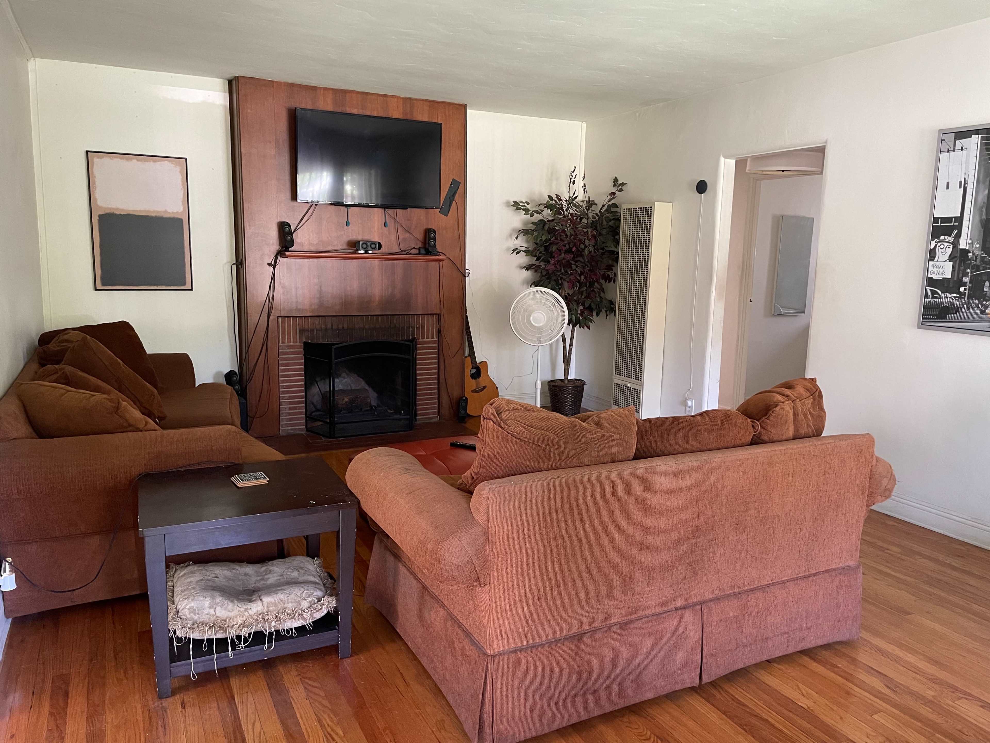 A living room with a sofa set, a coffee table, a wall-mounted TV, a fireplace, and a fan in front of a houseplant.