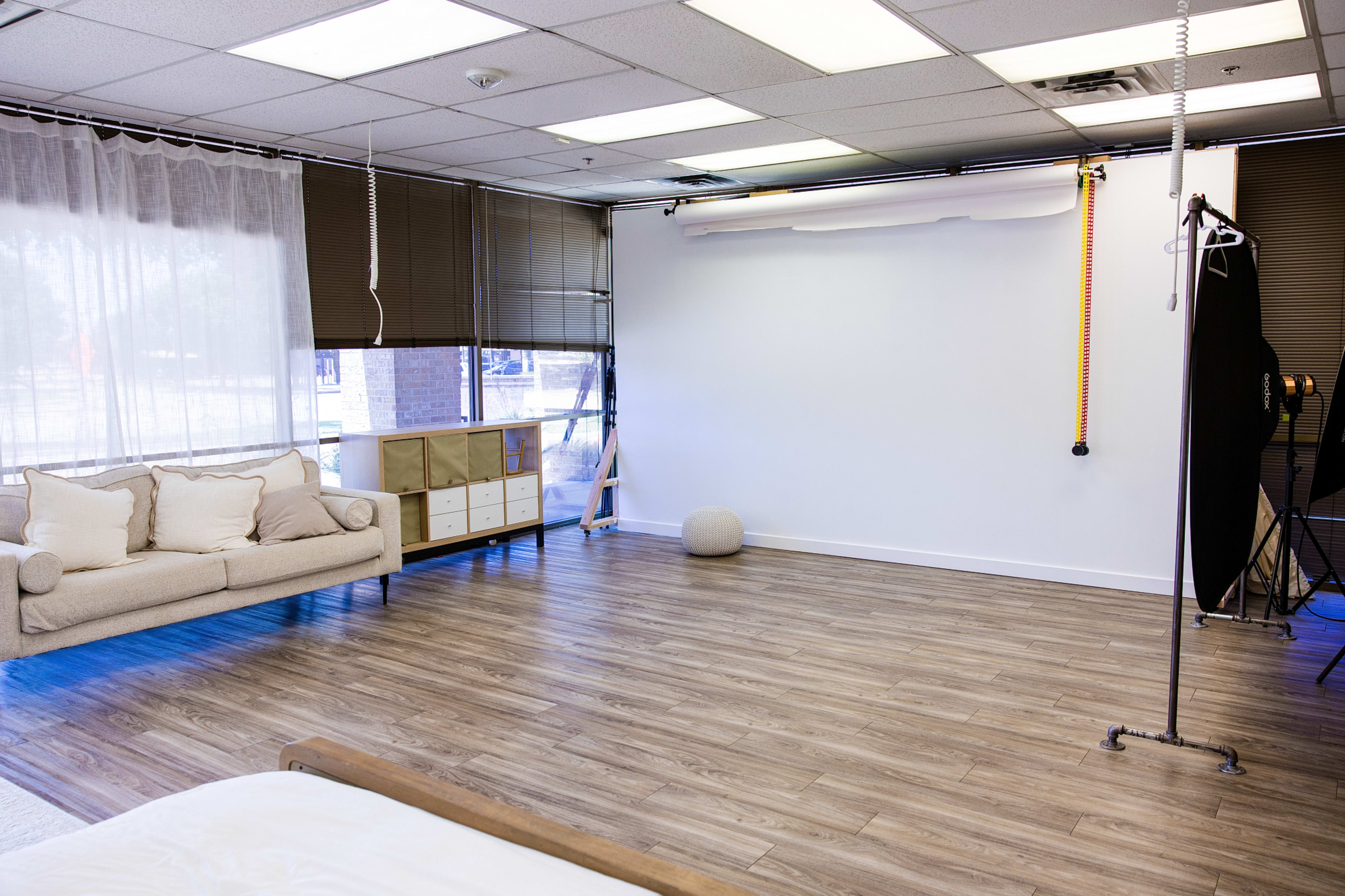 The image shows a spacious photography studio with a light-colored couch, a storage unit, and a large white backdrop arranged against a wall.