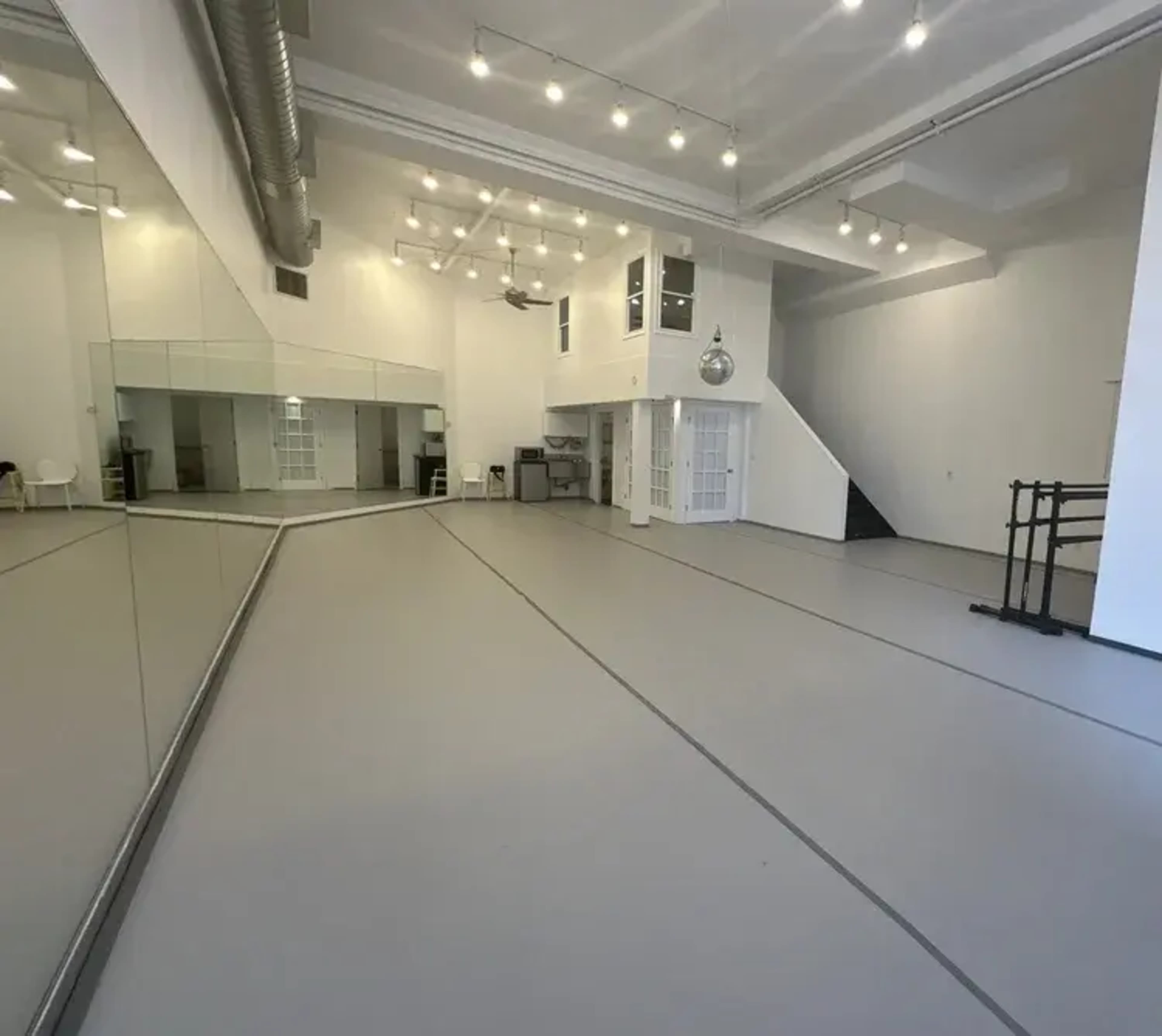 The image shows a spacious, well-lit dance studio with mirrors on one side and a minimalist design featuring a staircase and some furniture in the background.