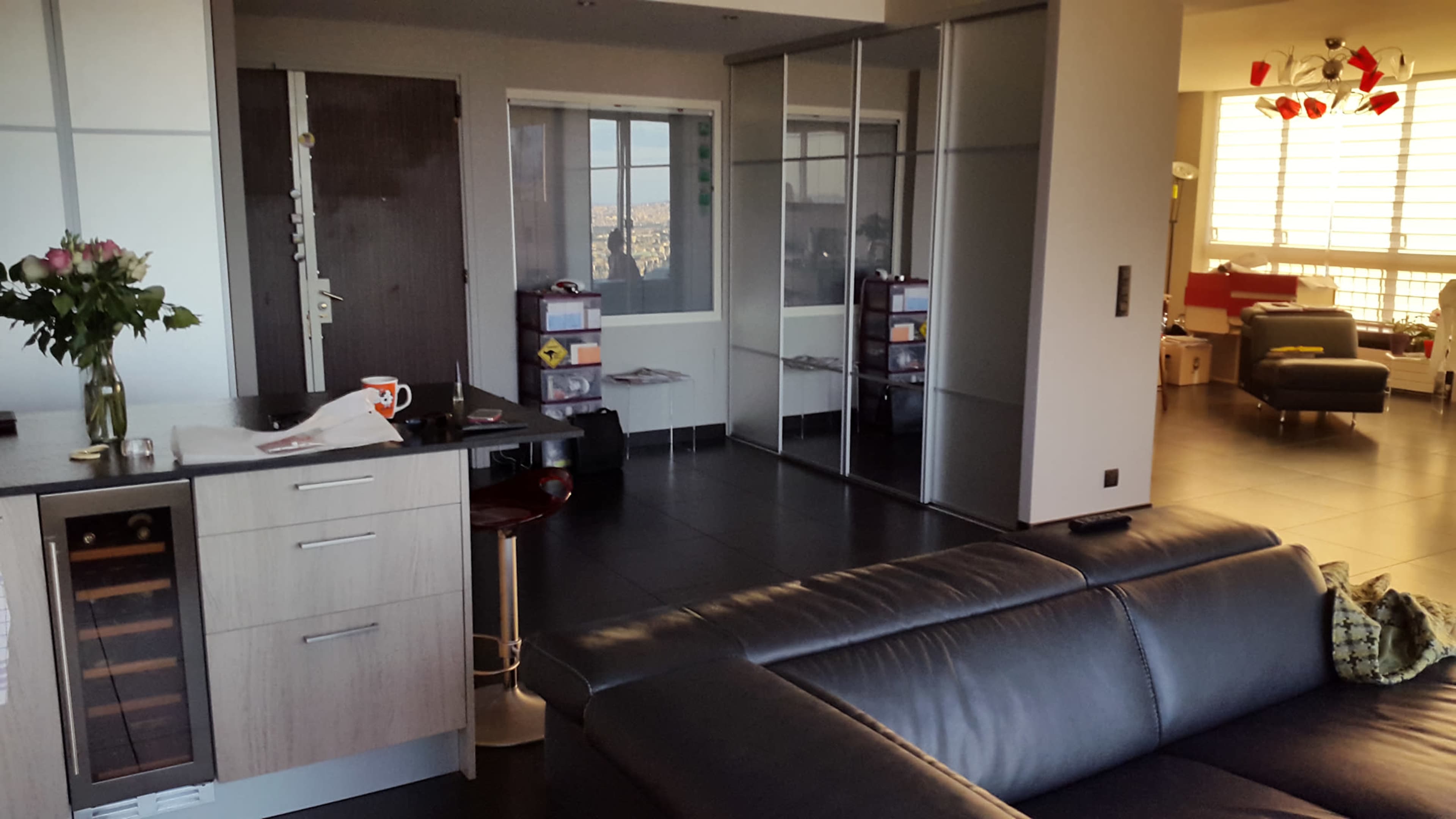 A modern apartment interior with a kitchen area, a living space featuring a black sofa, and glass sliding doors leading to a room with a view of the city.