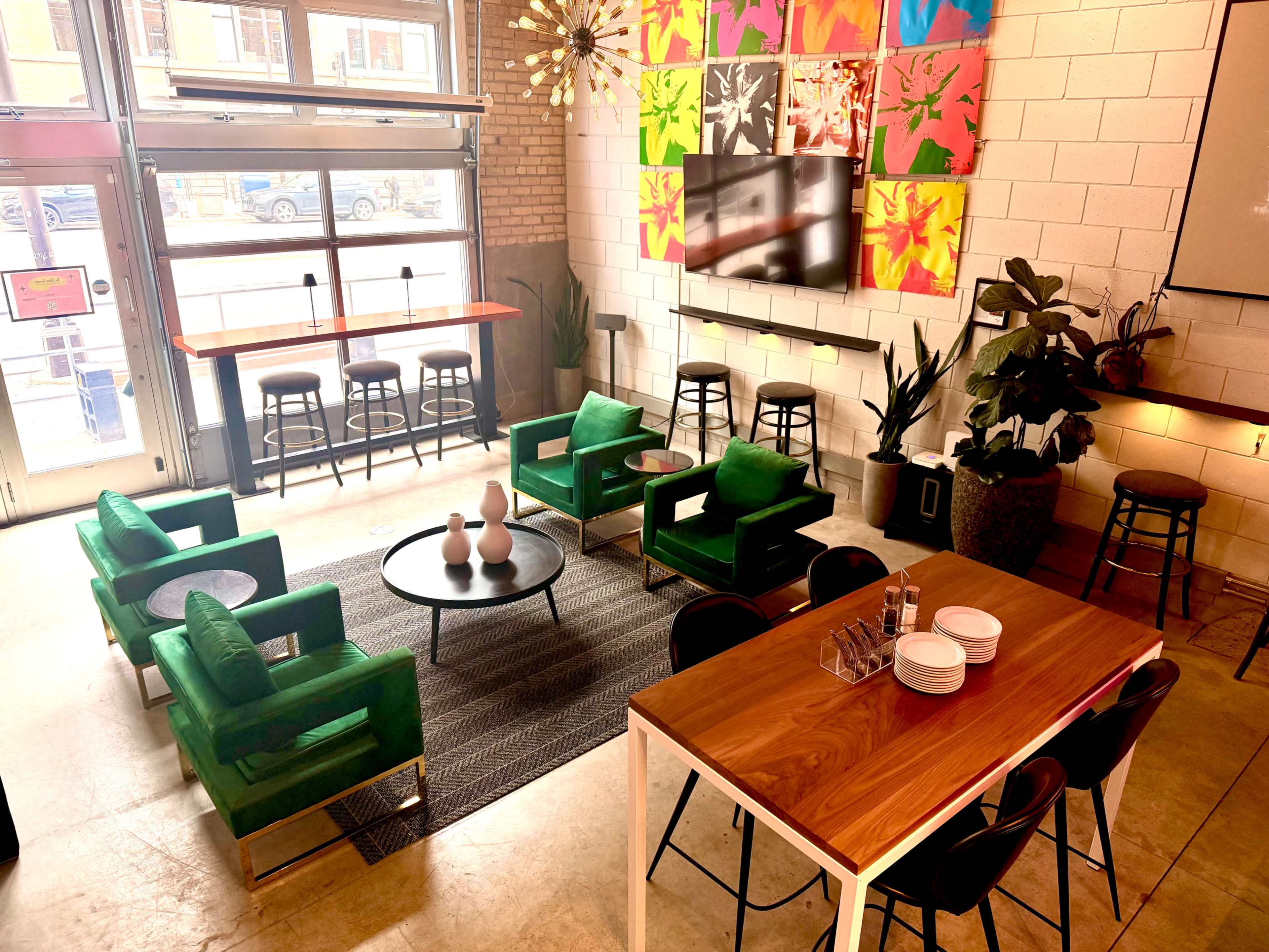 The image shows a modern cafe or lounge area with green chairs, a wooden dining table, and colorful wall art, featuring plants and a large TV mounted on the wall.