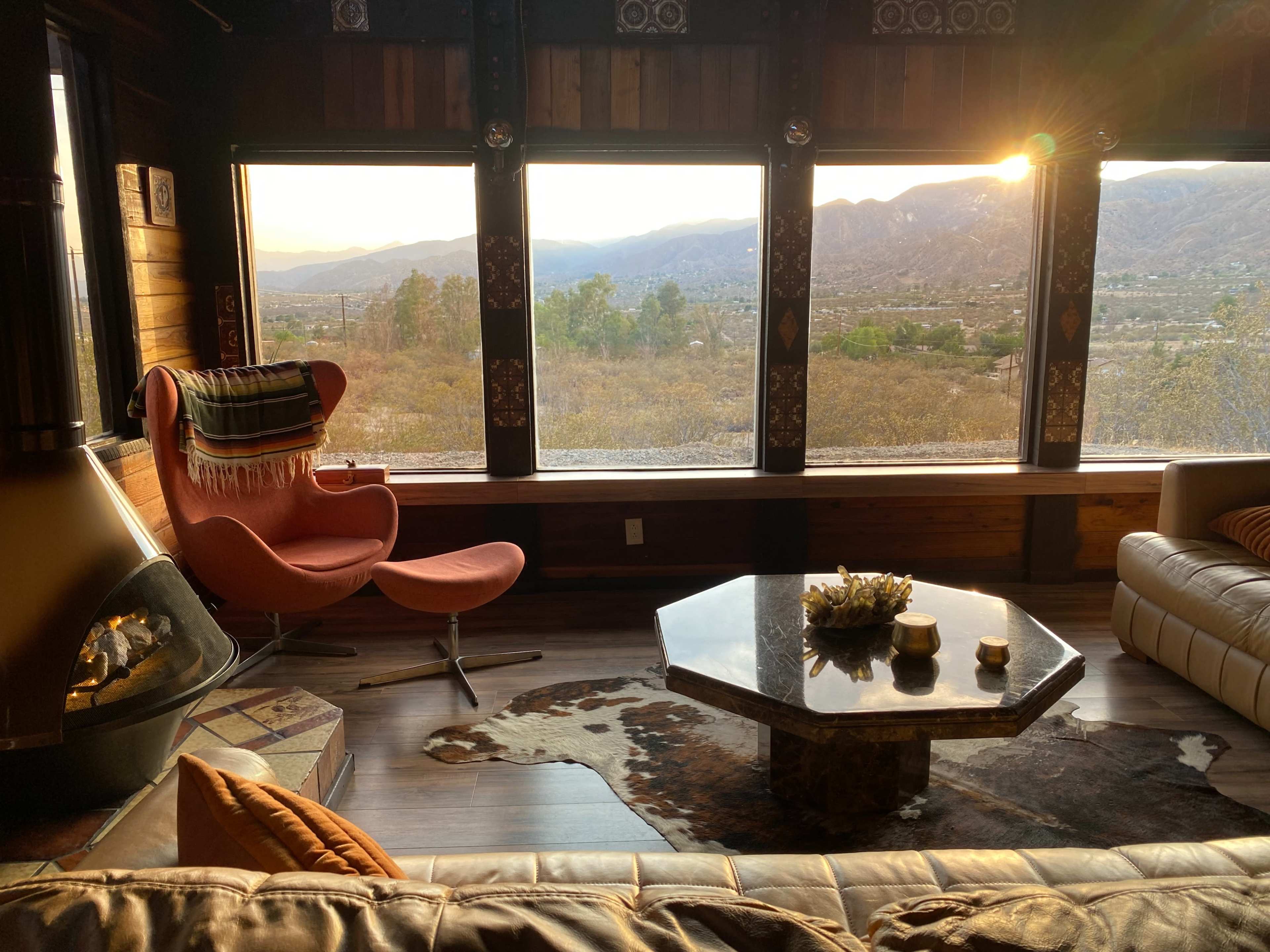 A cozy living room features large windows with a mountain view, an orange armchair, a stone coffee table, and a fireplace.