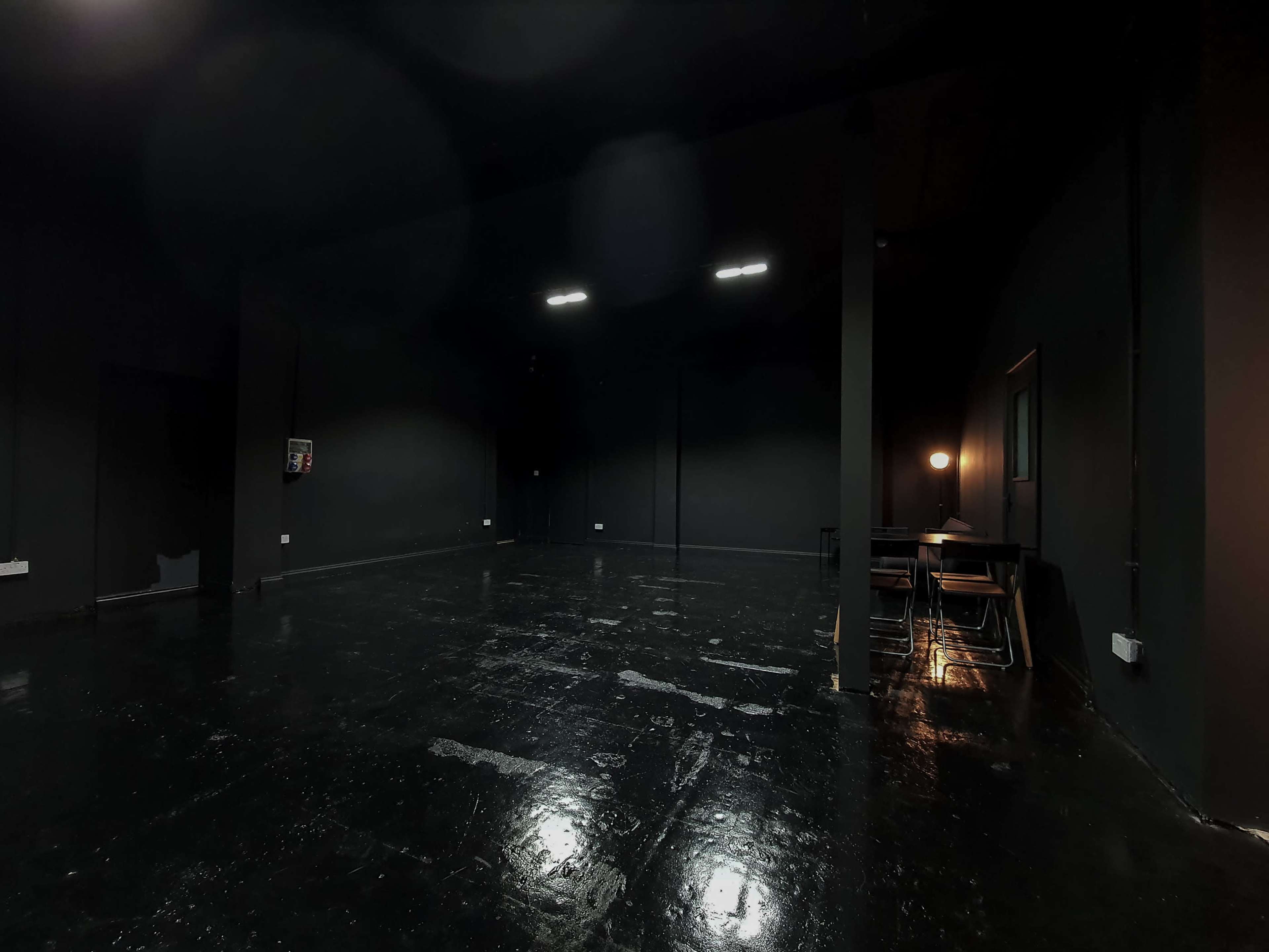 A spacious, dimly lit room with black walls and a shiny, reflective floor, featuring a small table and a lamp on one side.