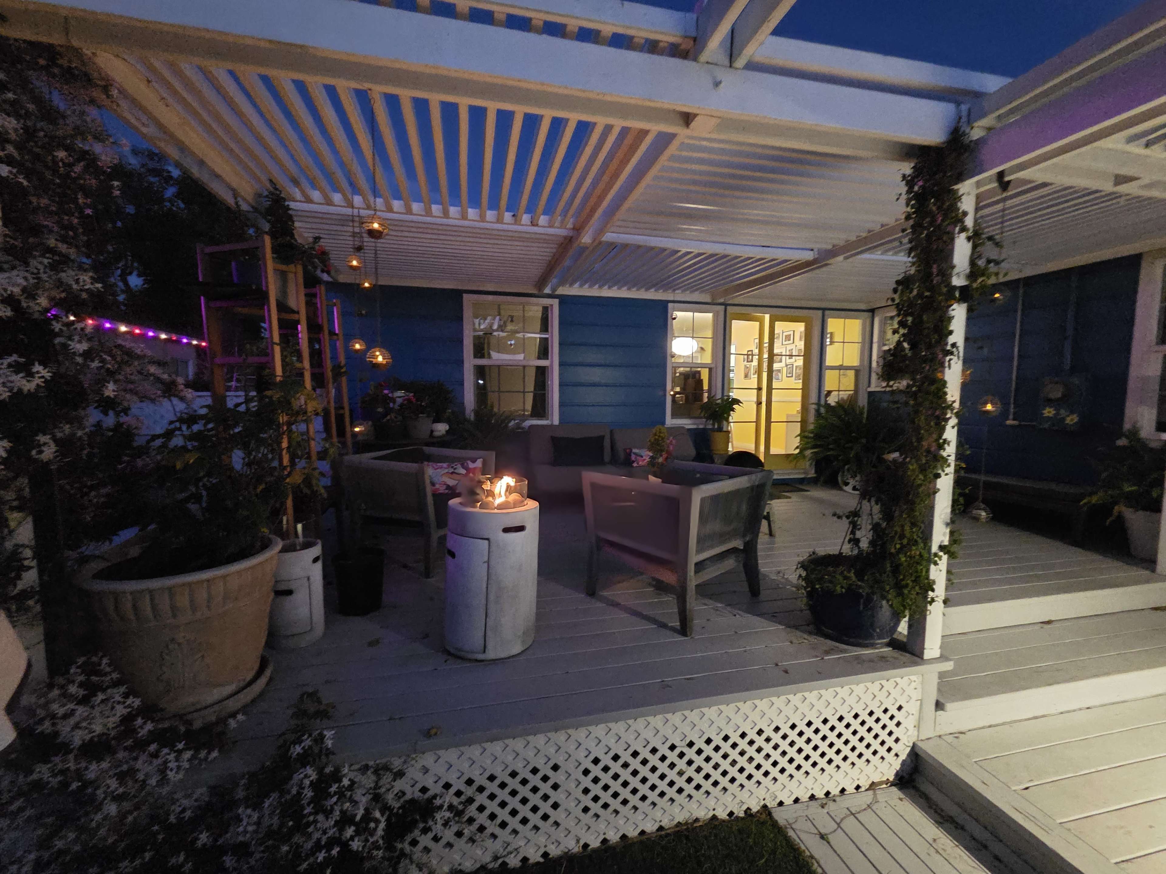 The image shows a cozy patio area with seating, potted plants, and decorative lighting, set against a blue house during twilight.