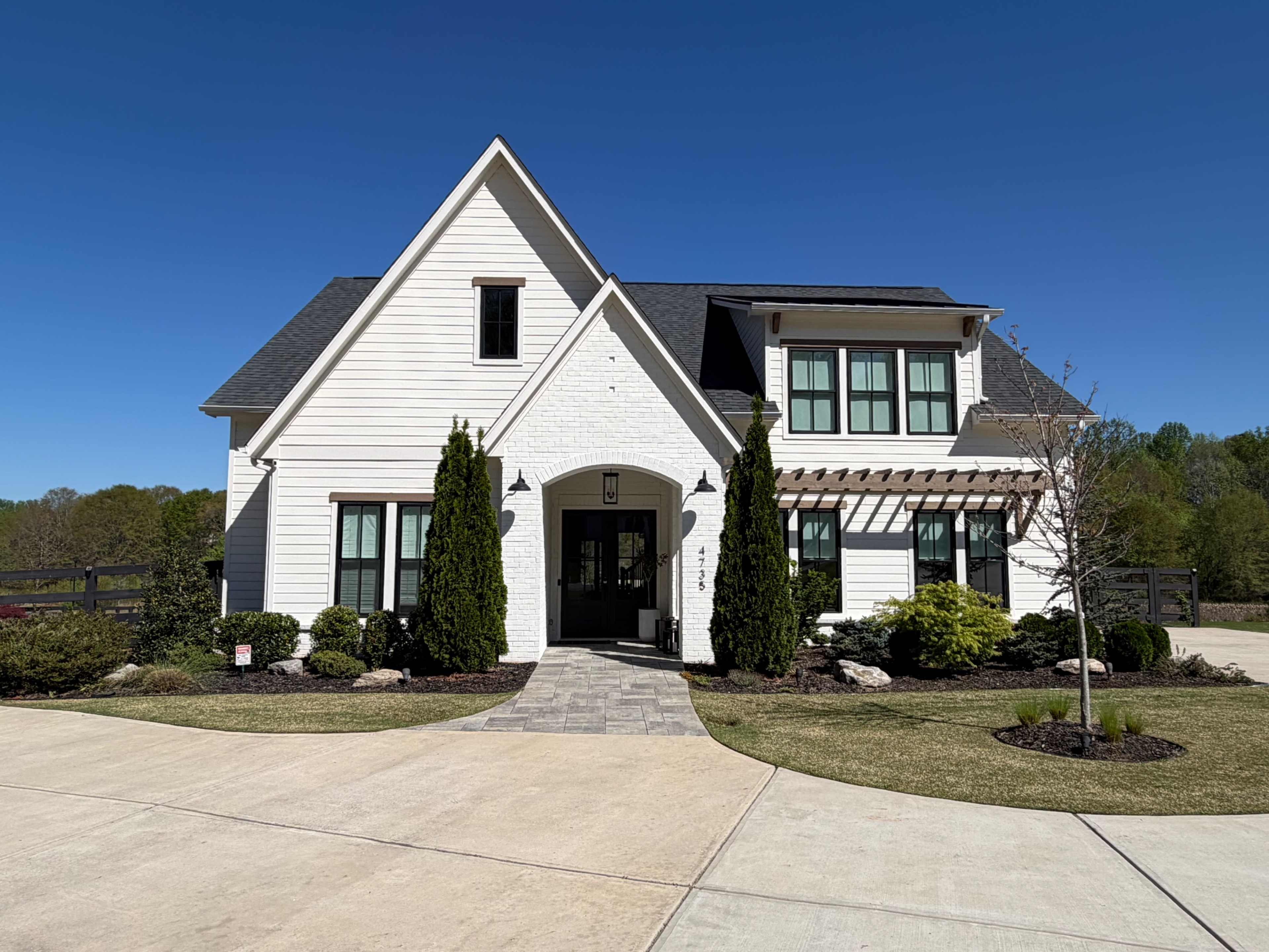 The image shows a modern white house with a black roof, green landscaping, and a paved driveway leading up to the entrance.