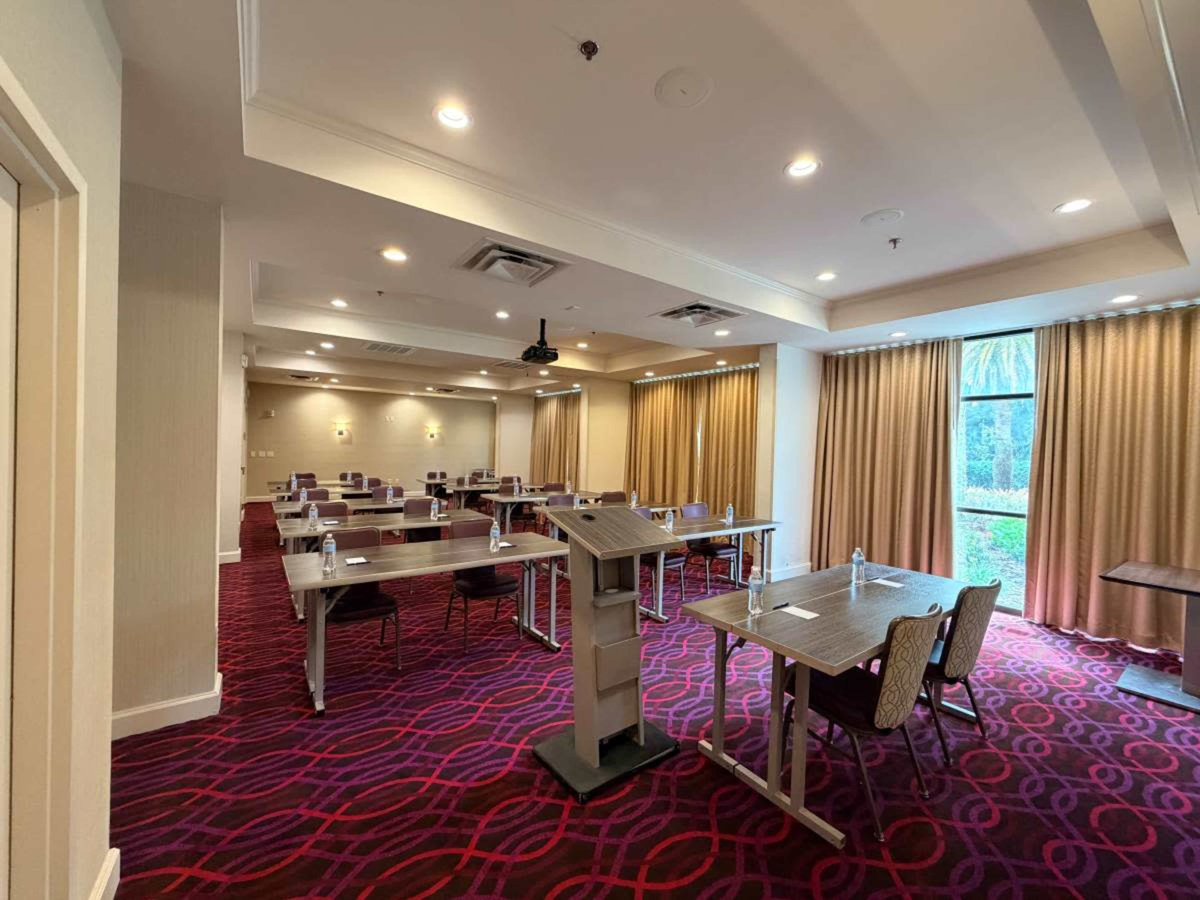 The image shows a spacious conference room set up with several tables and chairs, featuring a podium and large windows with curtains, allowing natural light to enter.