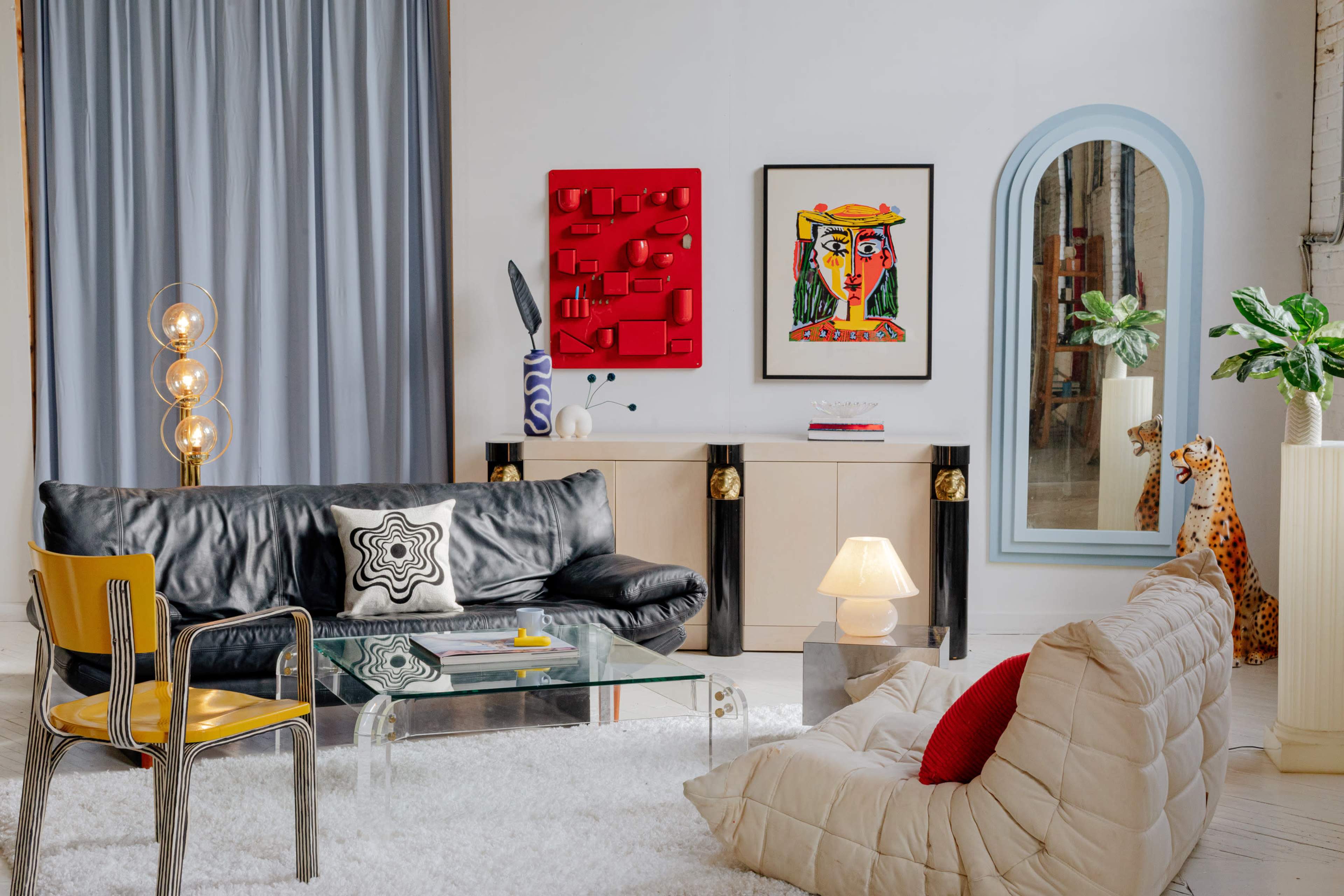 The image shows a stylish living room featuring a black leather sofa, a glass coffee table, colorful artwork on the walls, and modern decorative elements.