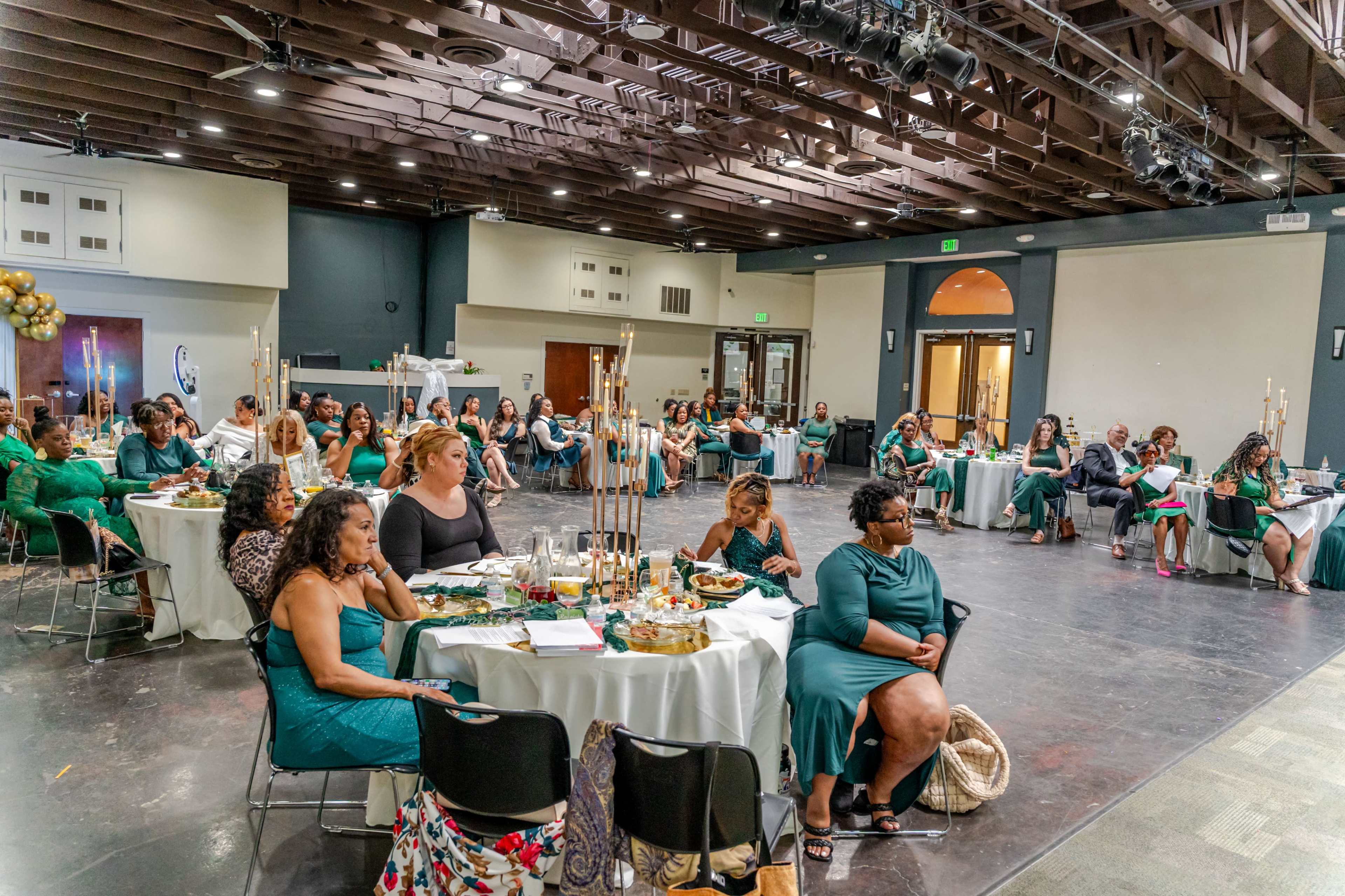 A large gathering of women in green attire is seated at round tables in a spacious hall, engaged in conversation and dining.