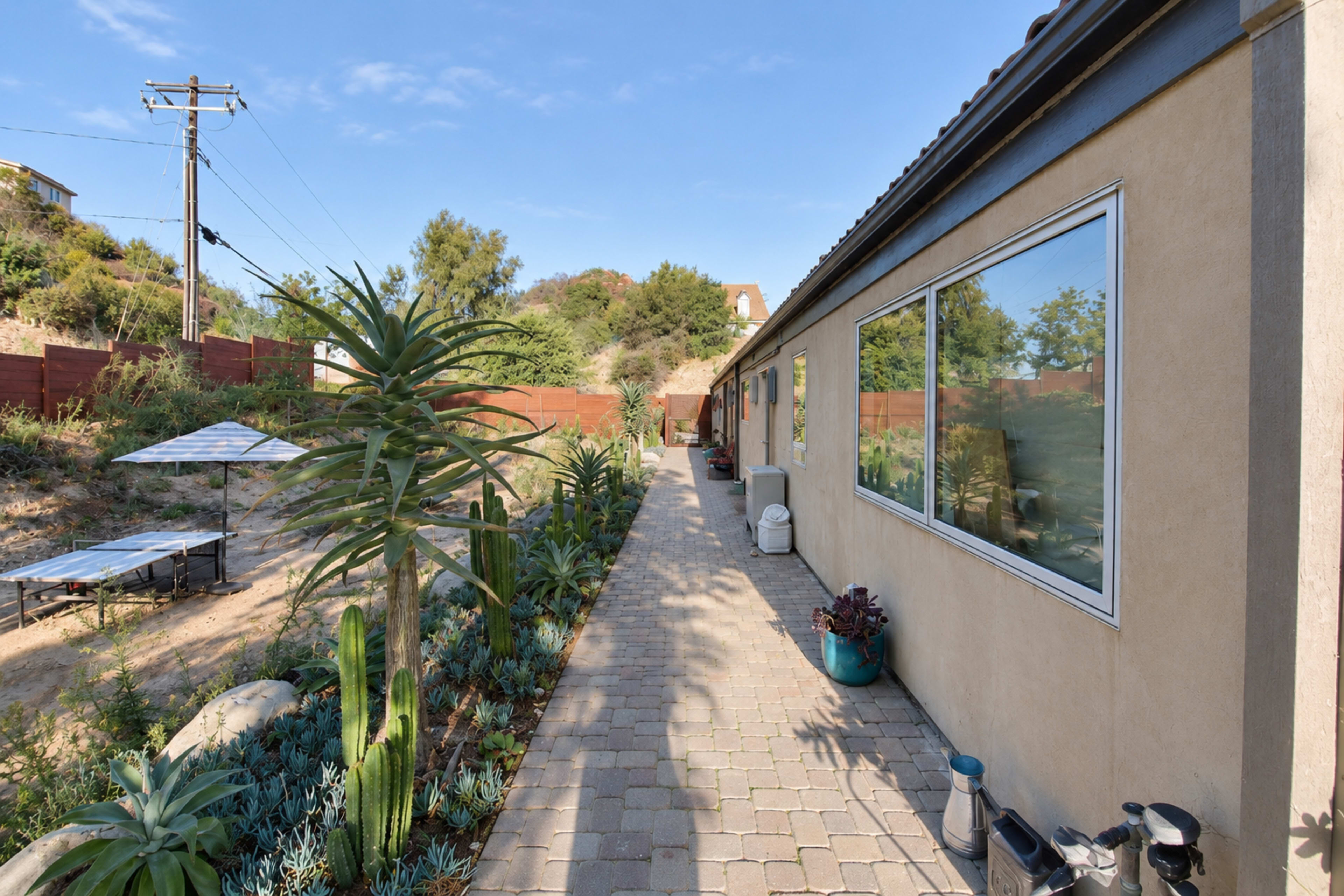 A narrow pathway lined with succulents leads alongside the exterior of a house with large windows, bordered by a green hillside and a wooden fence.