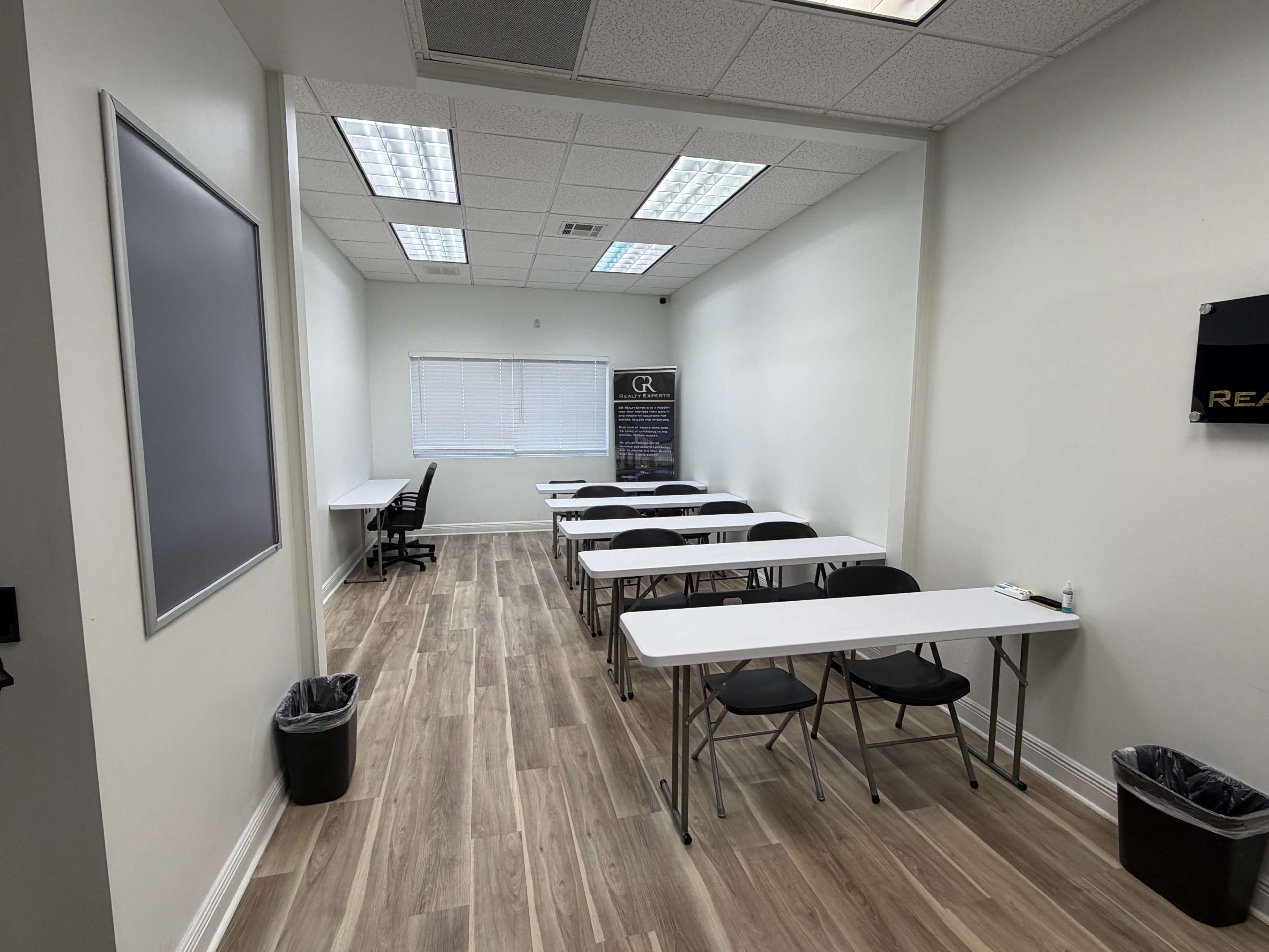 The image shows a well-lit classroom or meeting room with multiple tables and chairs arranged in rows, a whiteboard, and a window with blinds.