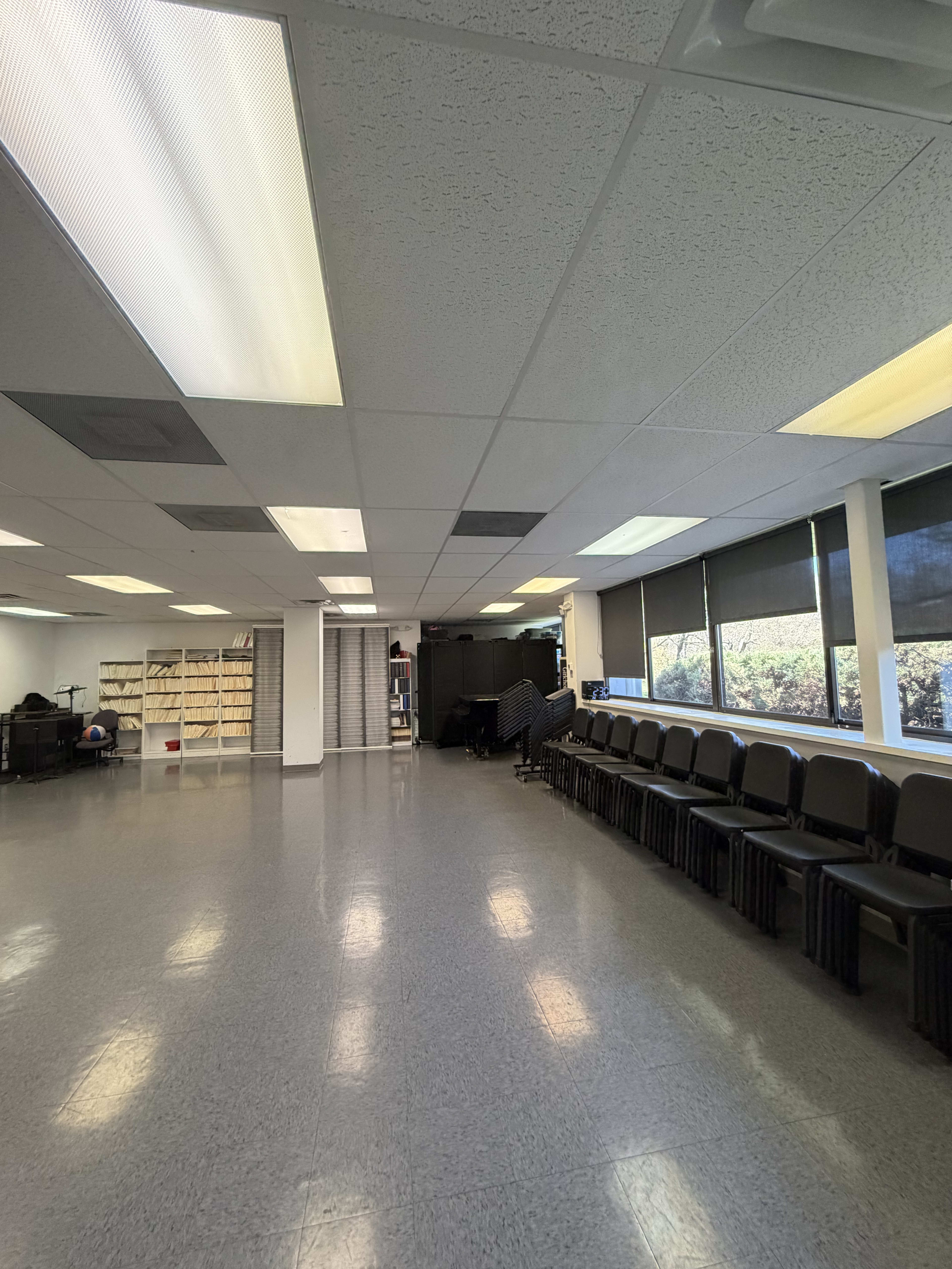 The image shows a spacious, well-lit room with a row of black chairs along one side, shelves stacked with items on another, and large windows allowing natural light to enter.