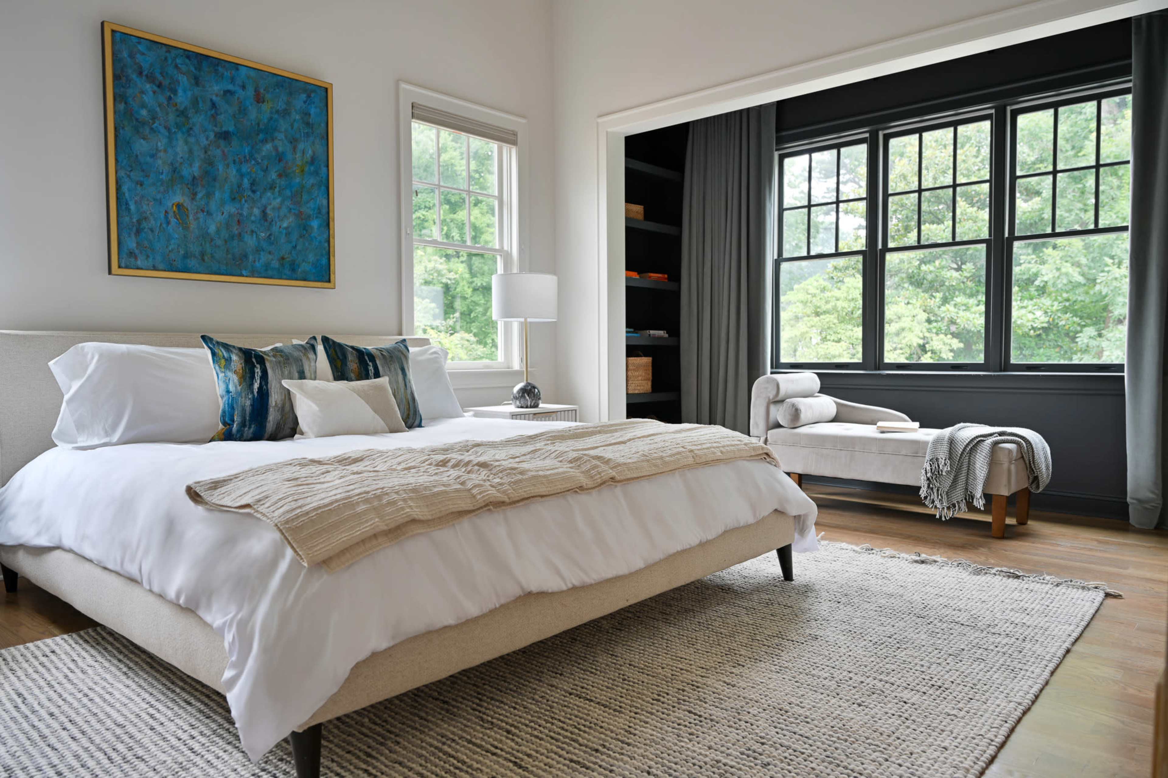 A modern bedroom with a neatly made bed, accented by pillows and a throw blanket, and a seating area by a large window.