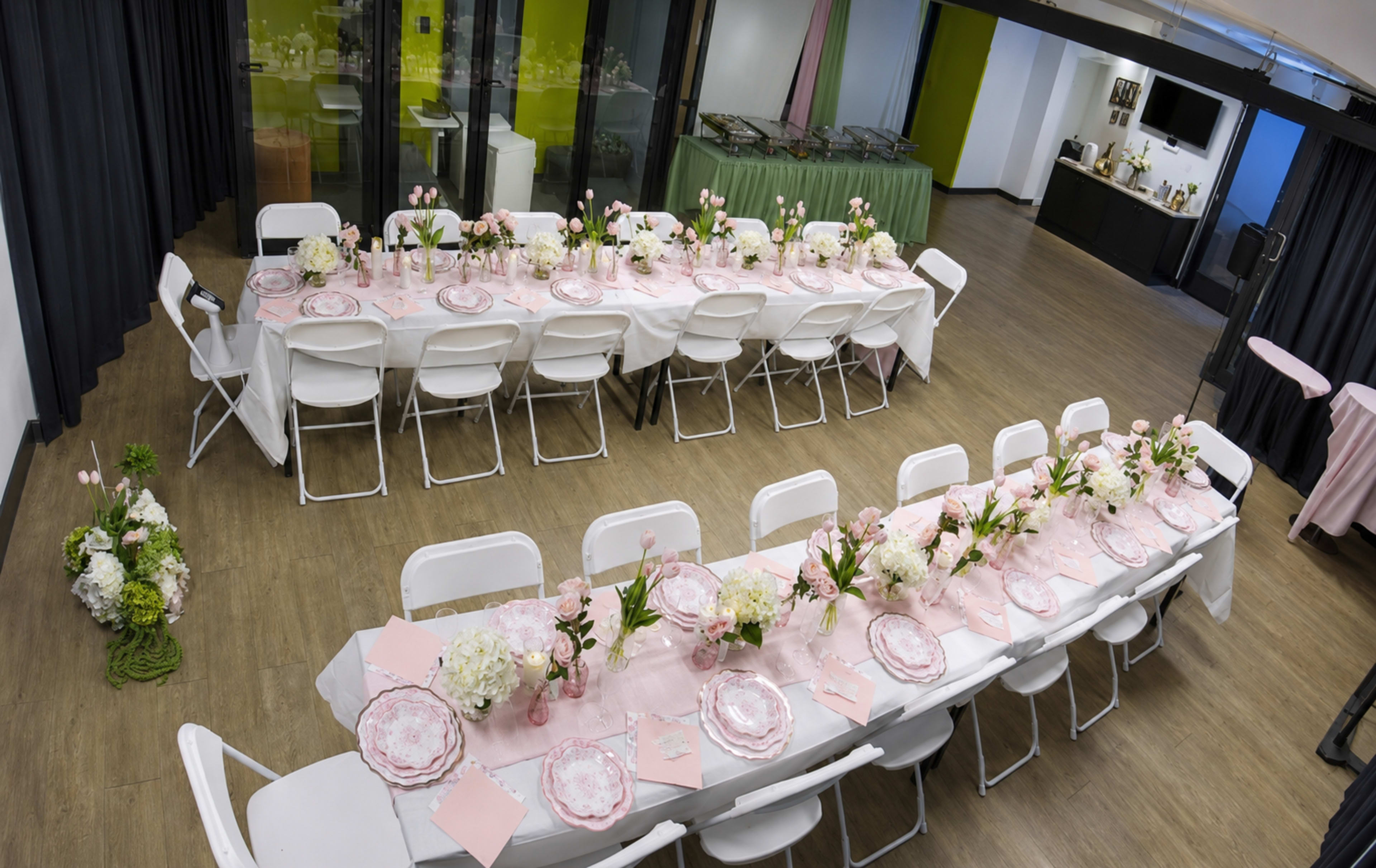 The image shows a large dining setup with multiple long tables elegantly arranged with pink tablecloths, floral centerpieces, and tableware in a modern indoor space.