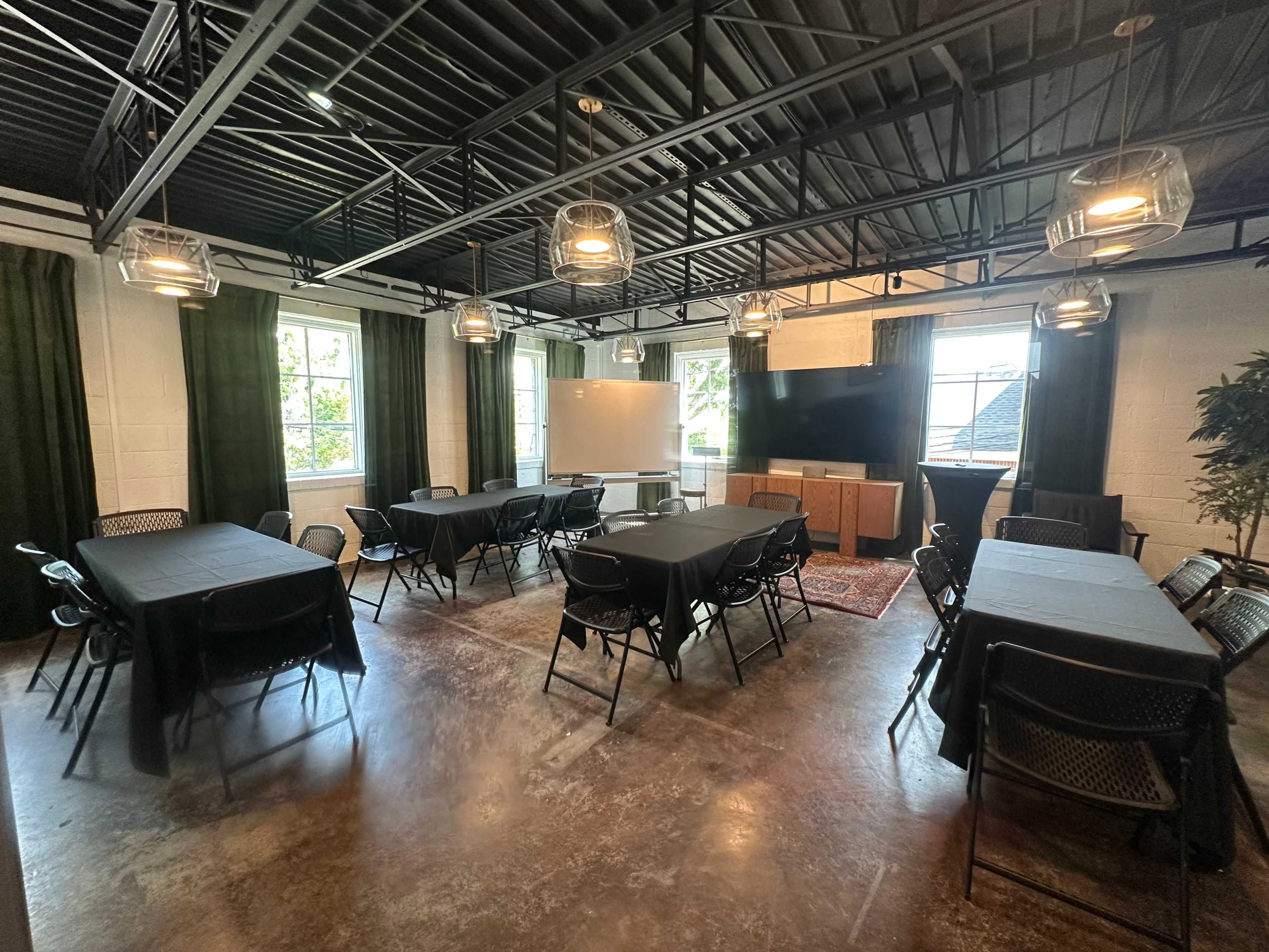 The image shows a conference room with multiple tables arranged in two clusters, surrounded by chairs, and featuring windows, a whiteboard, and a large screen.