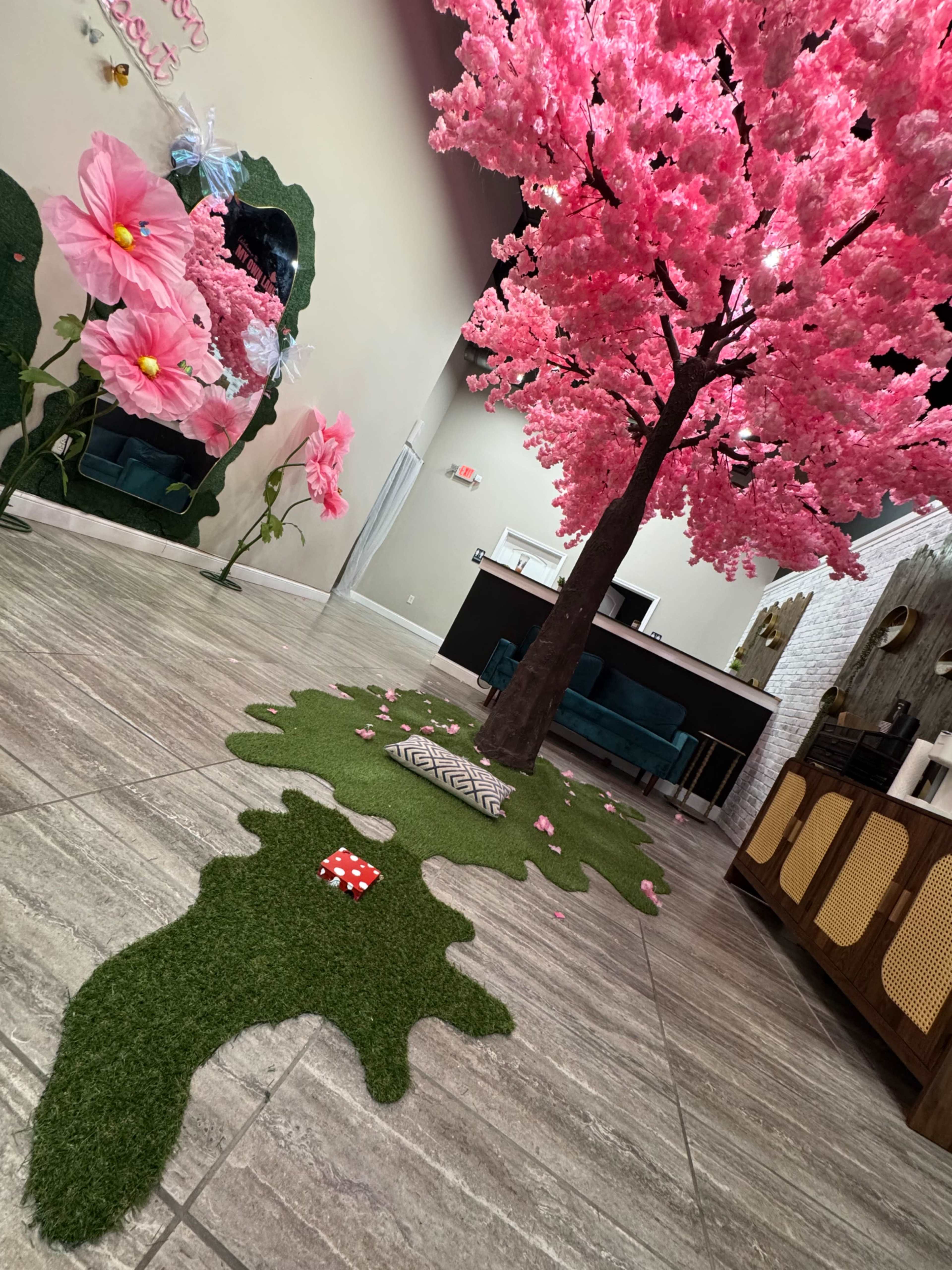 The image shows an indoor space featuring a large artificial pink cherry blossom tree with green grass-like flooring and decorative elements, including a small mushroom and patterned mat.