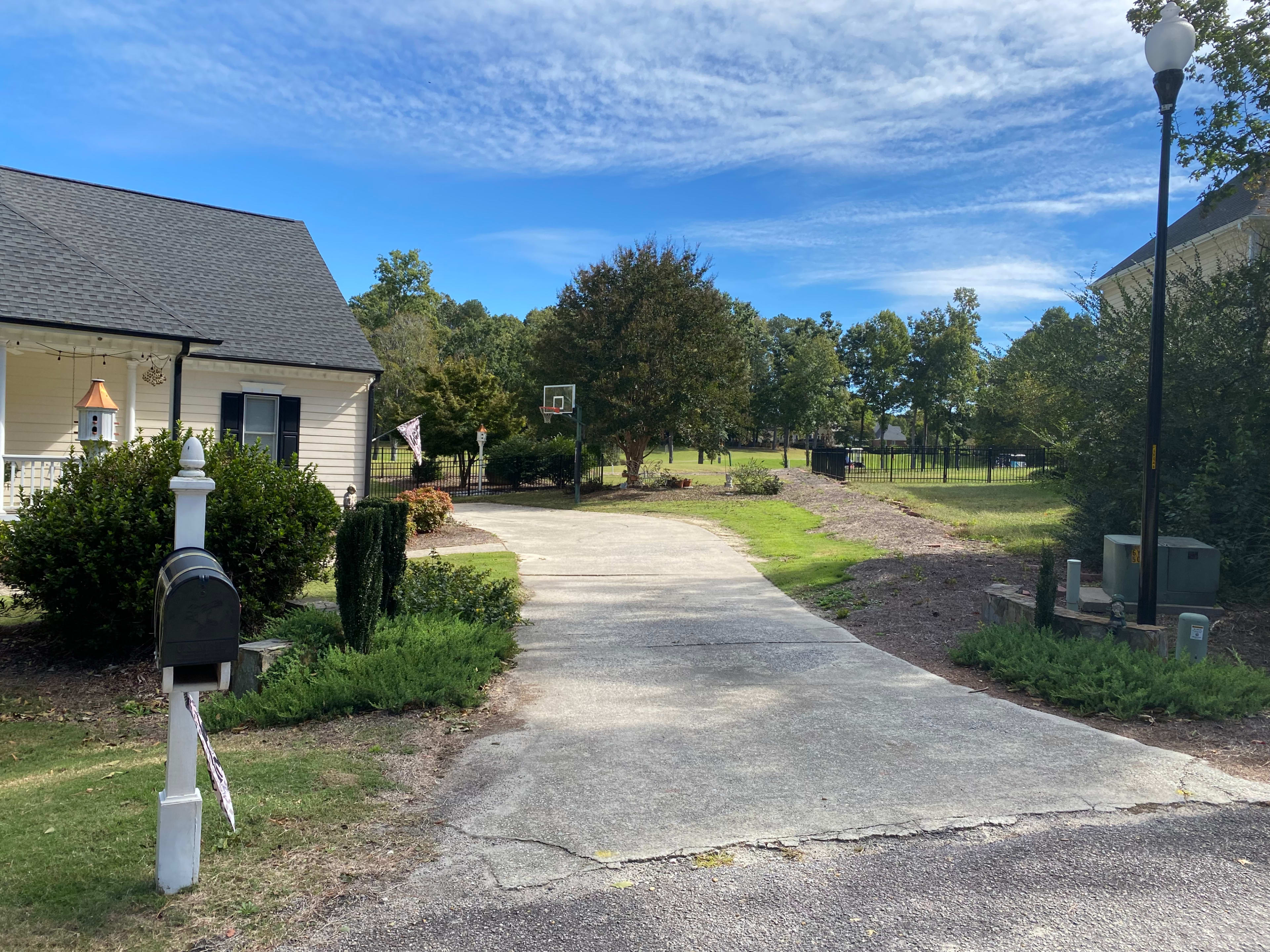 A paved driveway leads away from a house towards a basketball hoop and a grassy area.