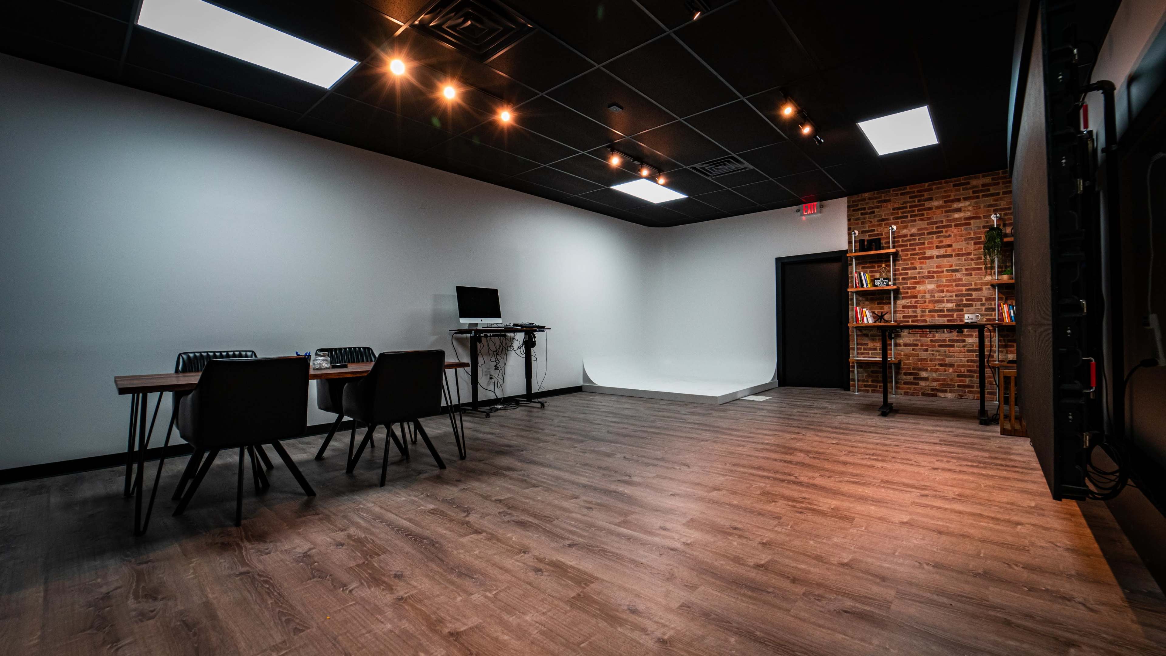 A modern, empty studio space with wooden flooring, two tables with chairs, a computer setup, and a brick wall with shelves.
