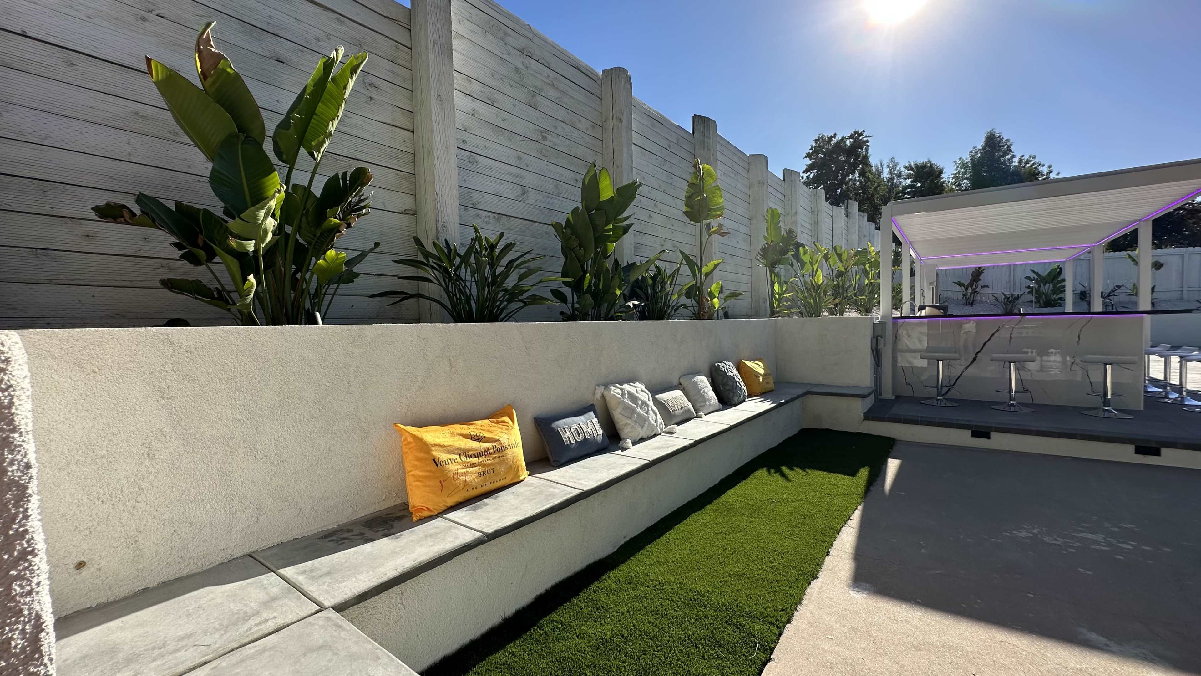 A low stone seating area adorned with cushions next to a green lawn, set against a backdrop of a concrete wall and tropical plants under a bright blue sky.
