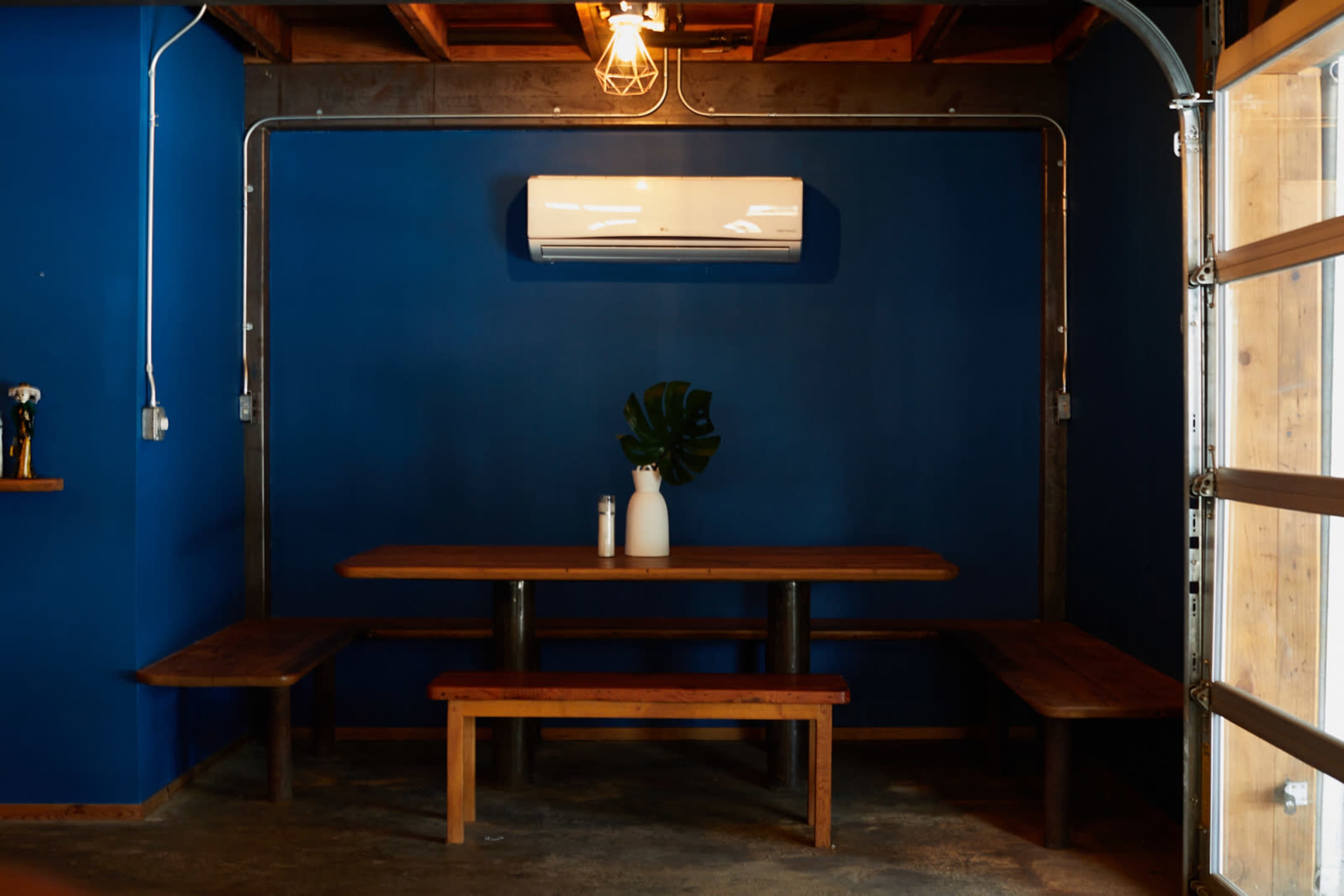 A wooden table with benches is positioned against a blue wall, topped with a vase and a bottle, under a ceiling light and an air conditioning unit.