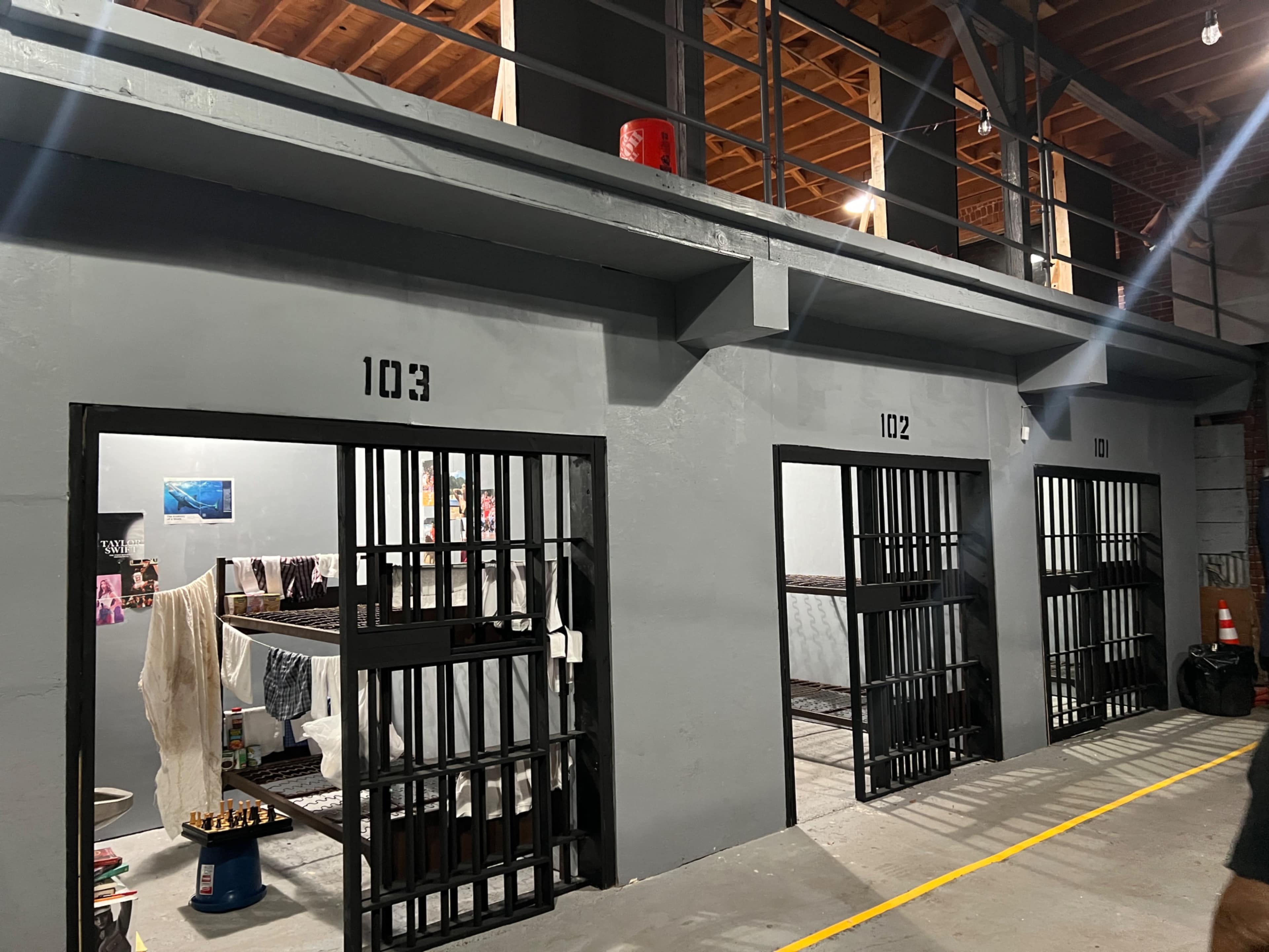 A section of a building with four barred cell doors, labeled 102, 103, and one unmarked, featuring bunk beds and a small table inside each space.