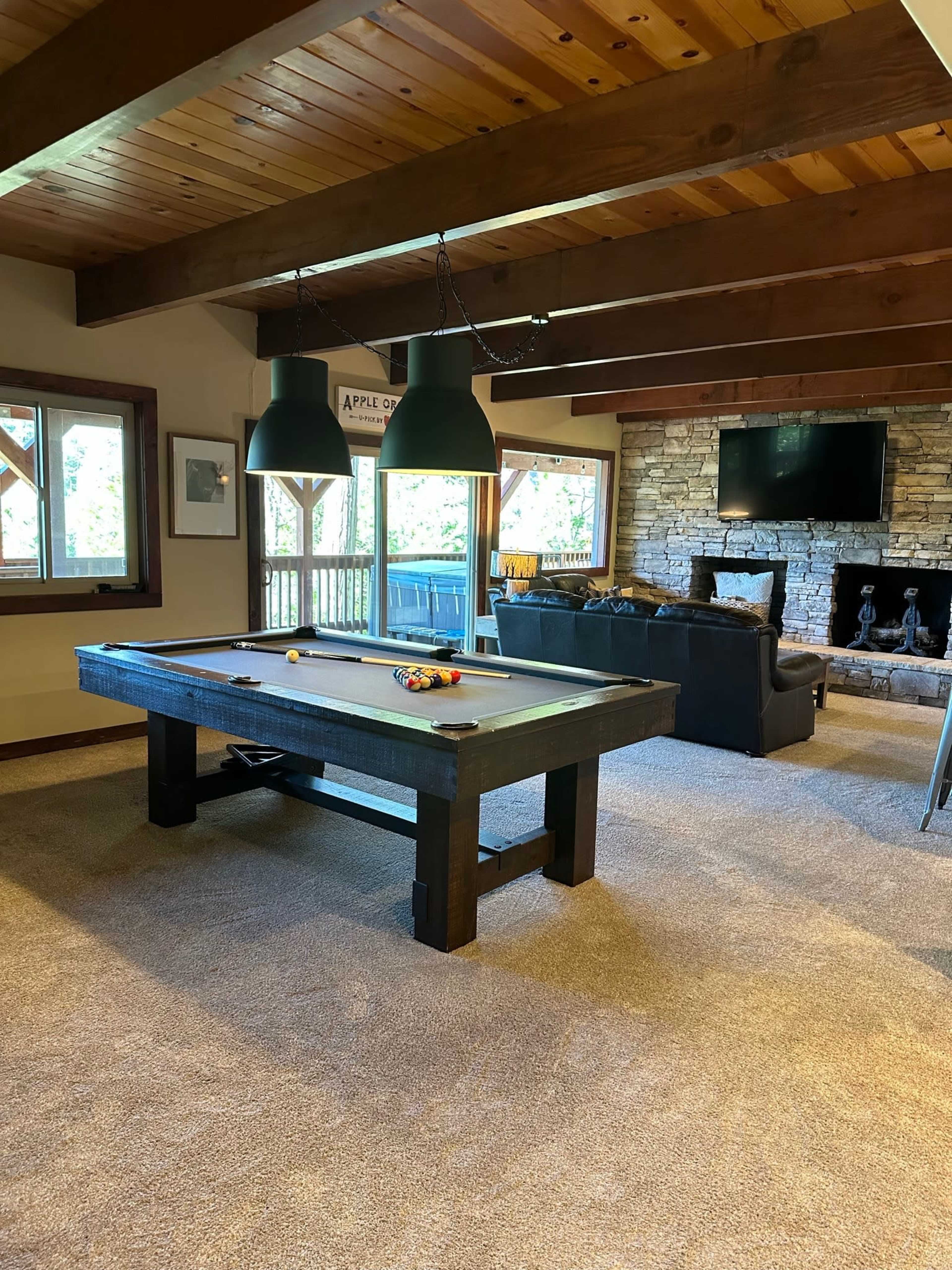 A spacious game room features a billiards table in the center, surrounded by windows and a stone fireplace.