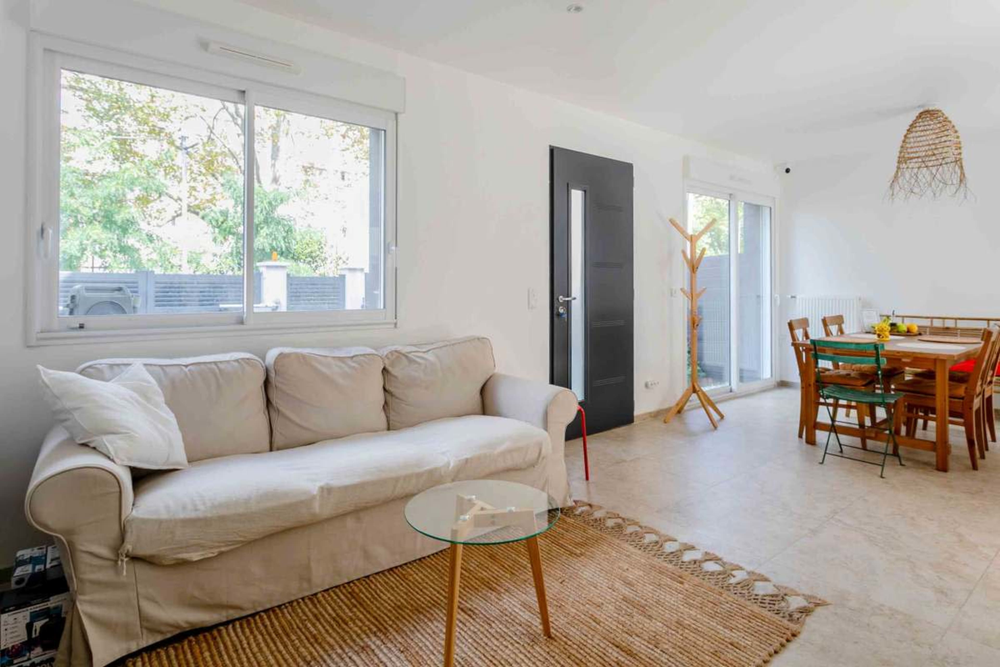 A light-filled living space features a beige couch, a glass coffee table, and a wooden dining table surrounded by chairs.