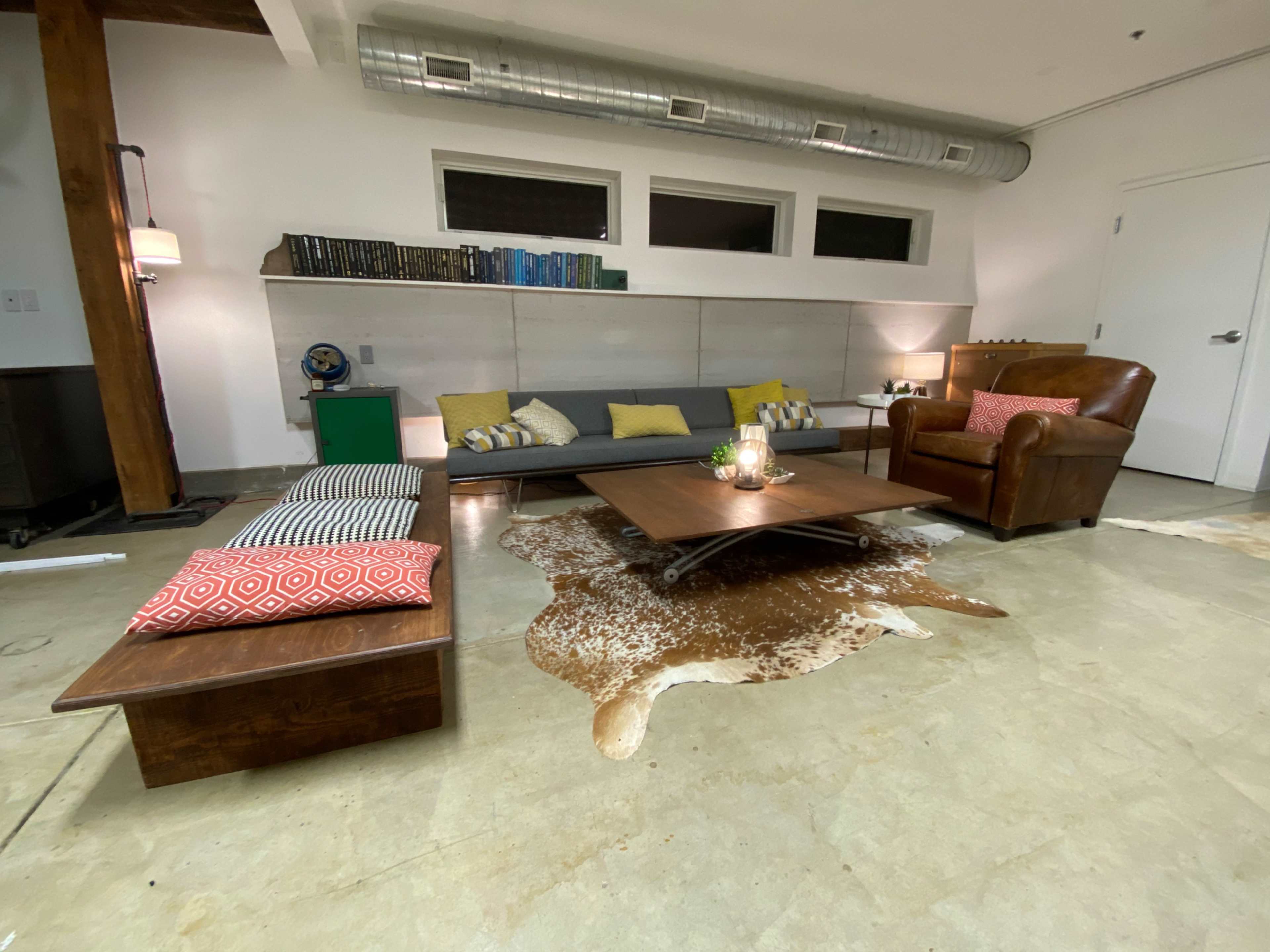 The image shows a modern living room featuring a gray sofa, a leather armchair, a wooden coffee table, and a cowhide rug on a concrete floor.