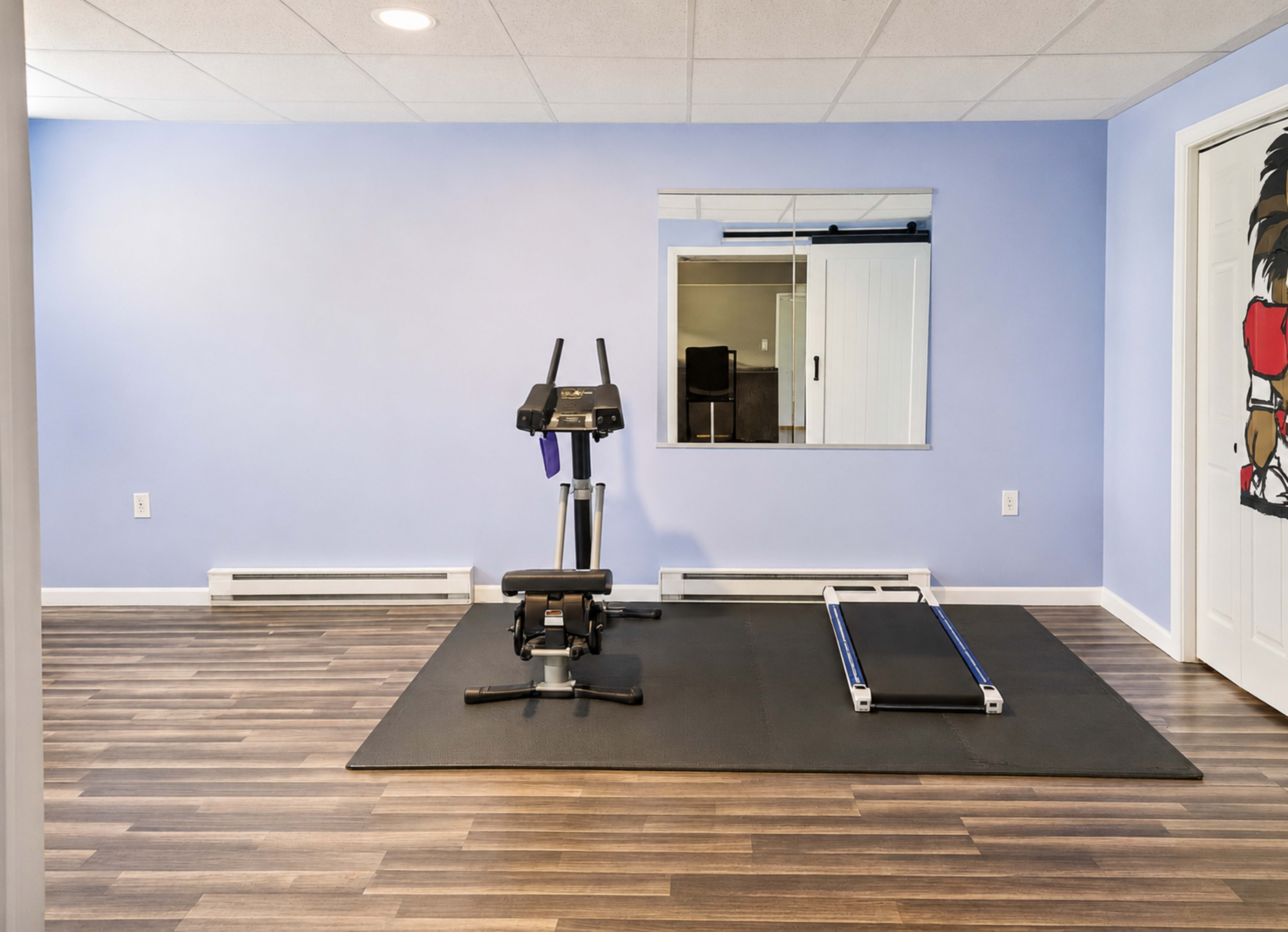 The image shows a home gym with exercise equipment, including a rowing machine and a flat exercise mat, set against a light blue wall and wooden flooring.