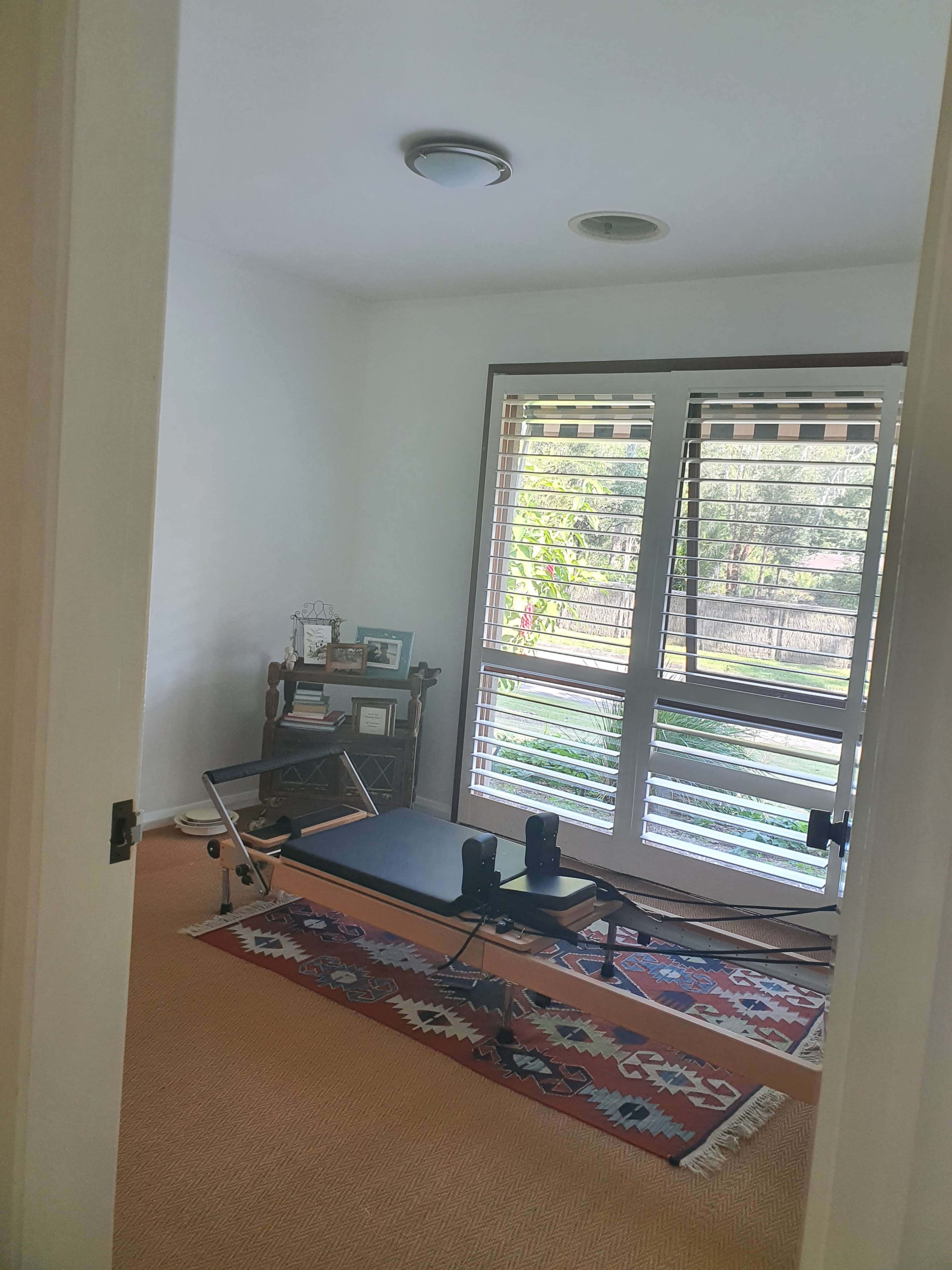 A small room with a reformer machine for Pilates, a rug on the floor, and windows with blinds overlooking greenery.