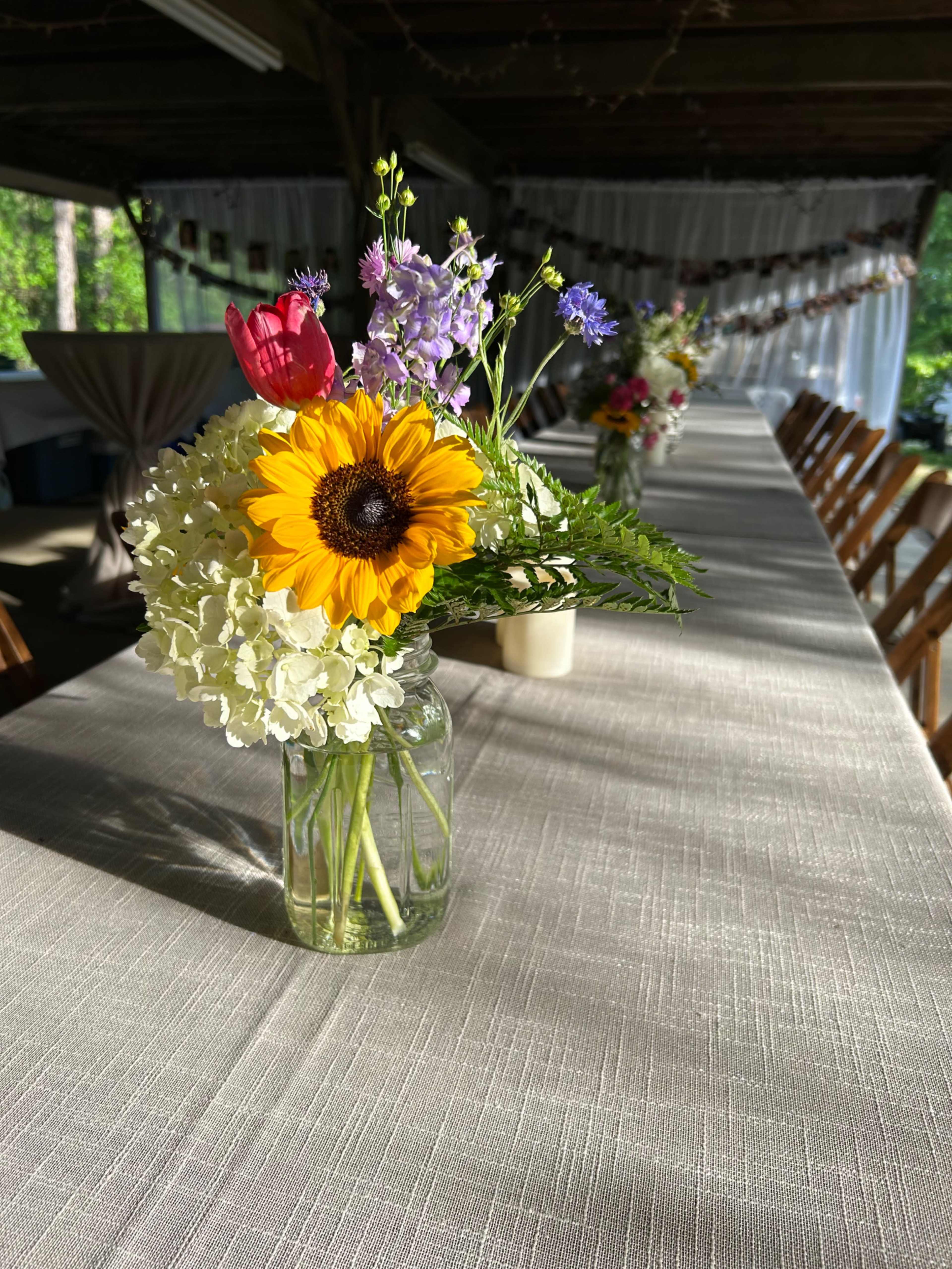 A jar of flowers, including a sunflower and various blooms, sits on a table covered with a textured linen cloth in a decorated outdoor space.