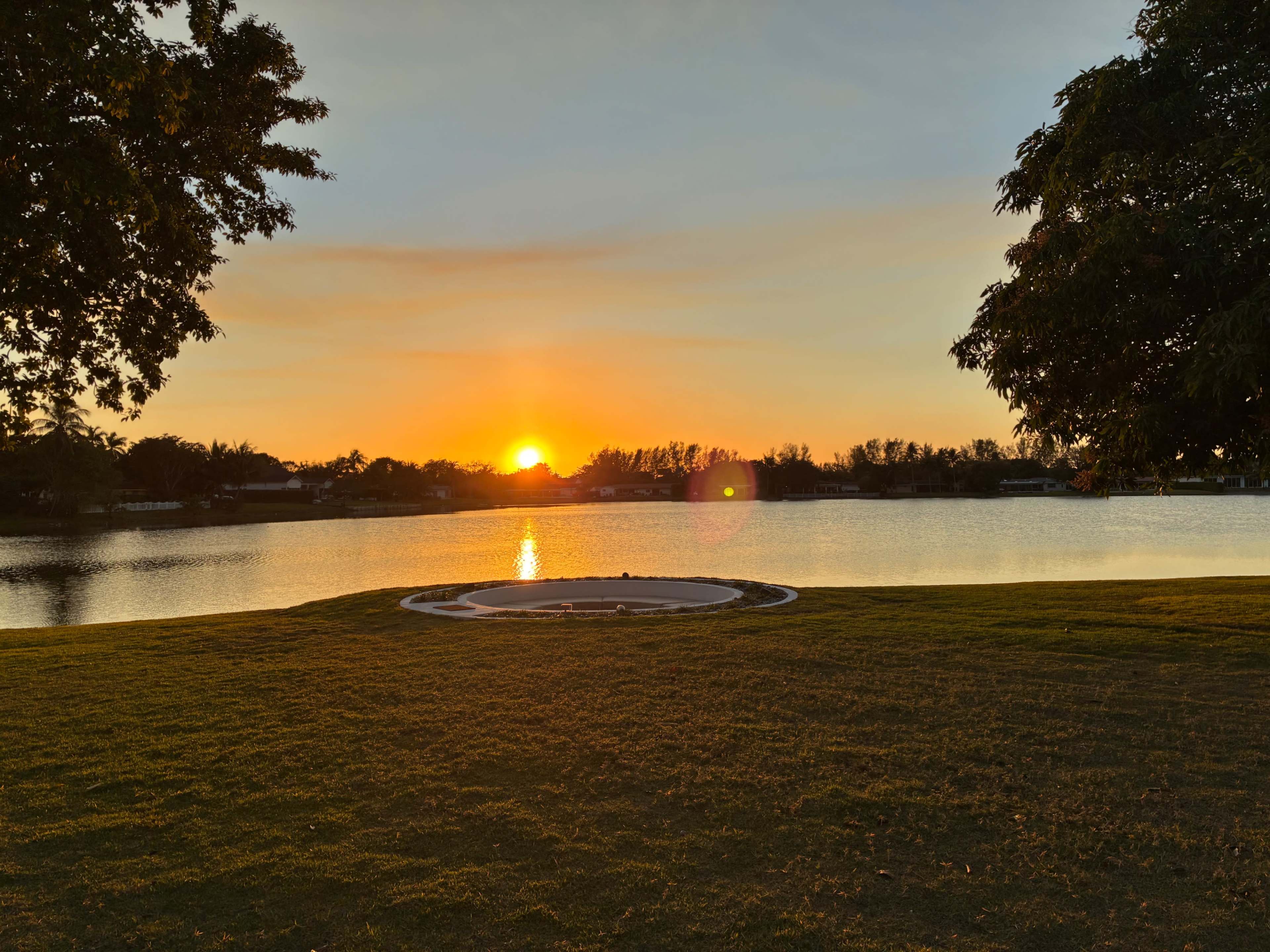 The sun sets over a tranquil lake, casting warm light on the surrounding trees and grassy area.